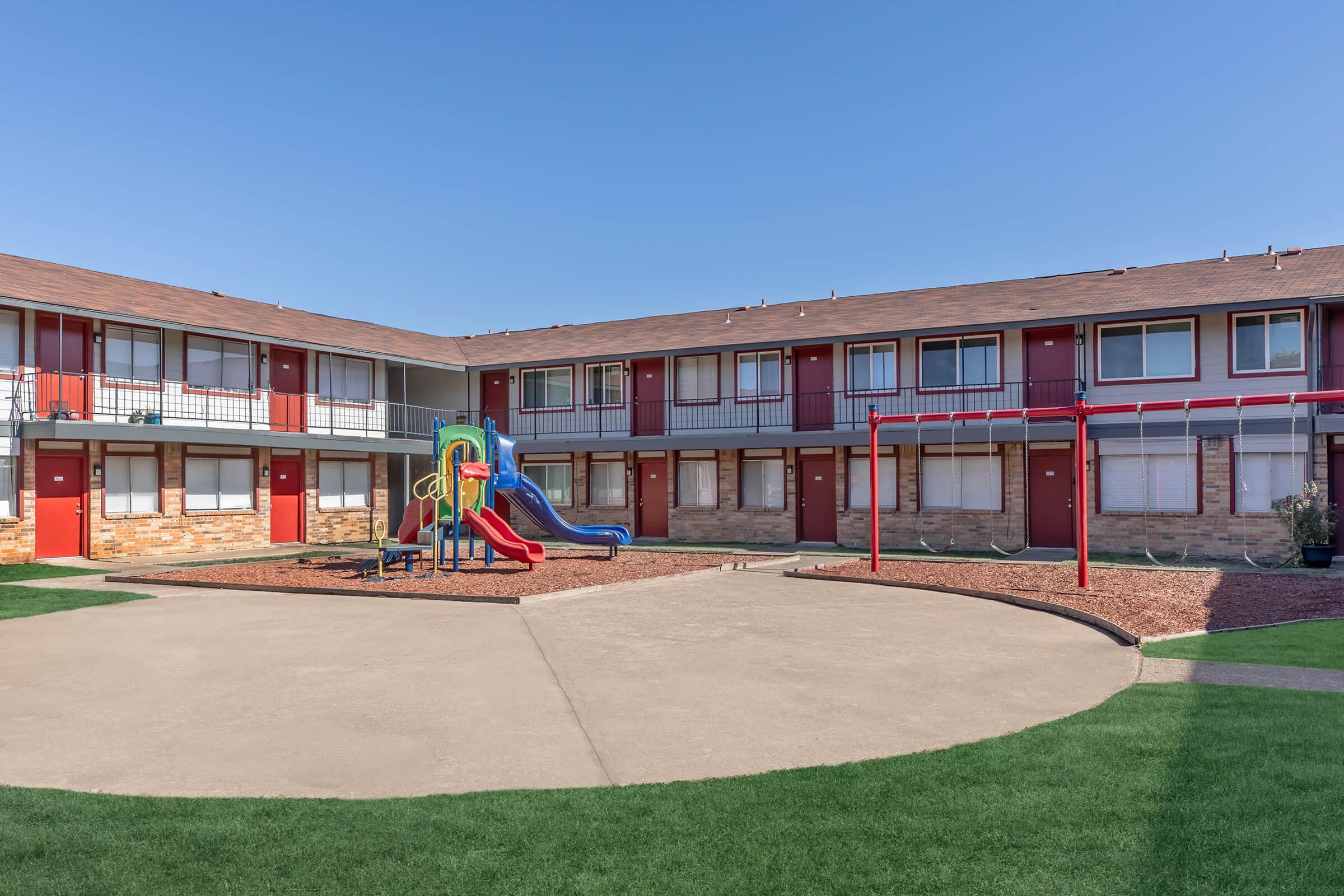 A courtyard of a two-story apartment complex featuring red doors and balconies. In the center, there is a playground with a slide and swings, surrounded by landscaped grass and gravel. The sky is clear and blue, creating a bright and inviting atmosphere.