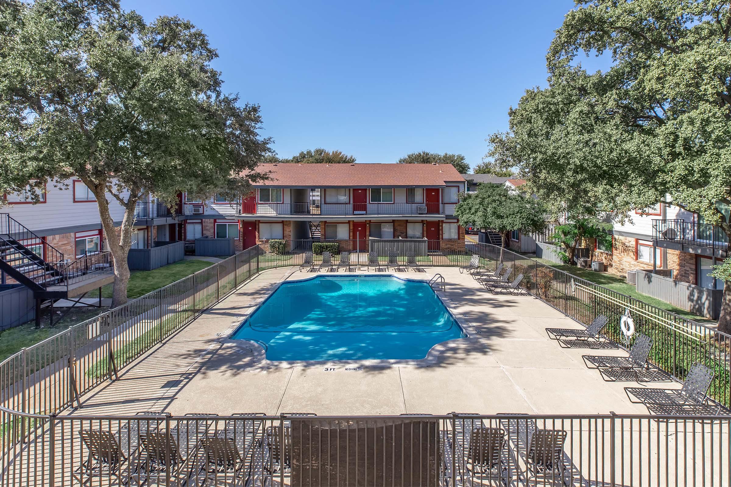 A swimming pool surrounded by lounge chairs, situated in the courtyard of an apartment complex. The area features trees and two-story buildings with red doors in the background, clear blue sky overhead. Fenced pool area with a clean, inviting atmosphere.