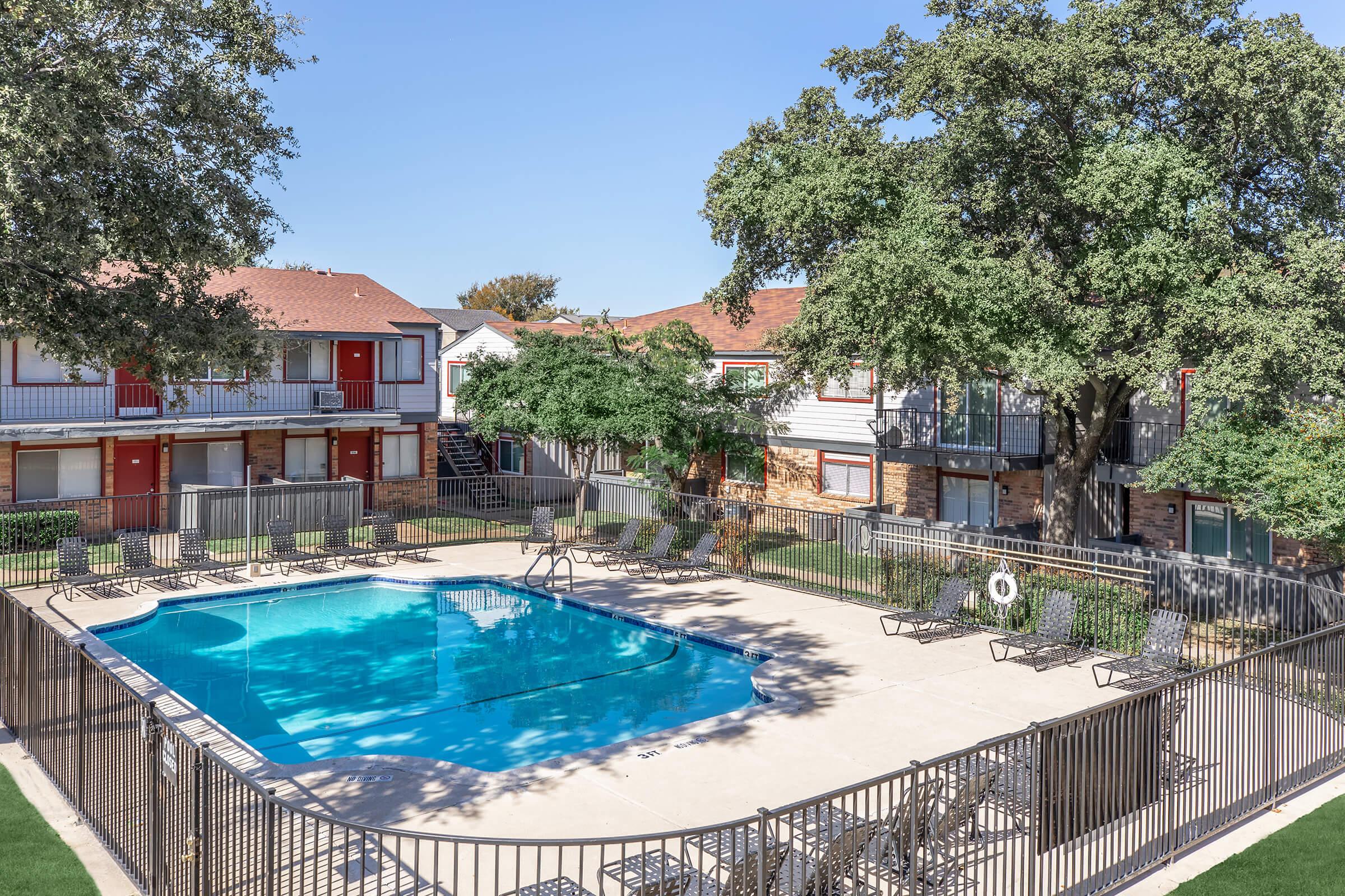 A clear blue swimming pool surrounded by a wrought iron fence, with lounge chairs arranged around it. In the background, there are apartment buildings with red doors and lush trees providing shade, under a clear blue sky.