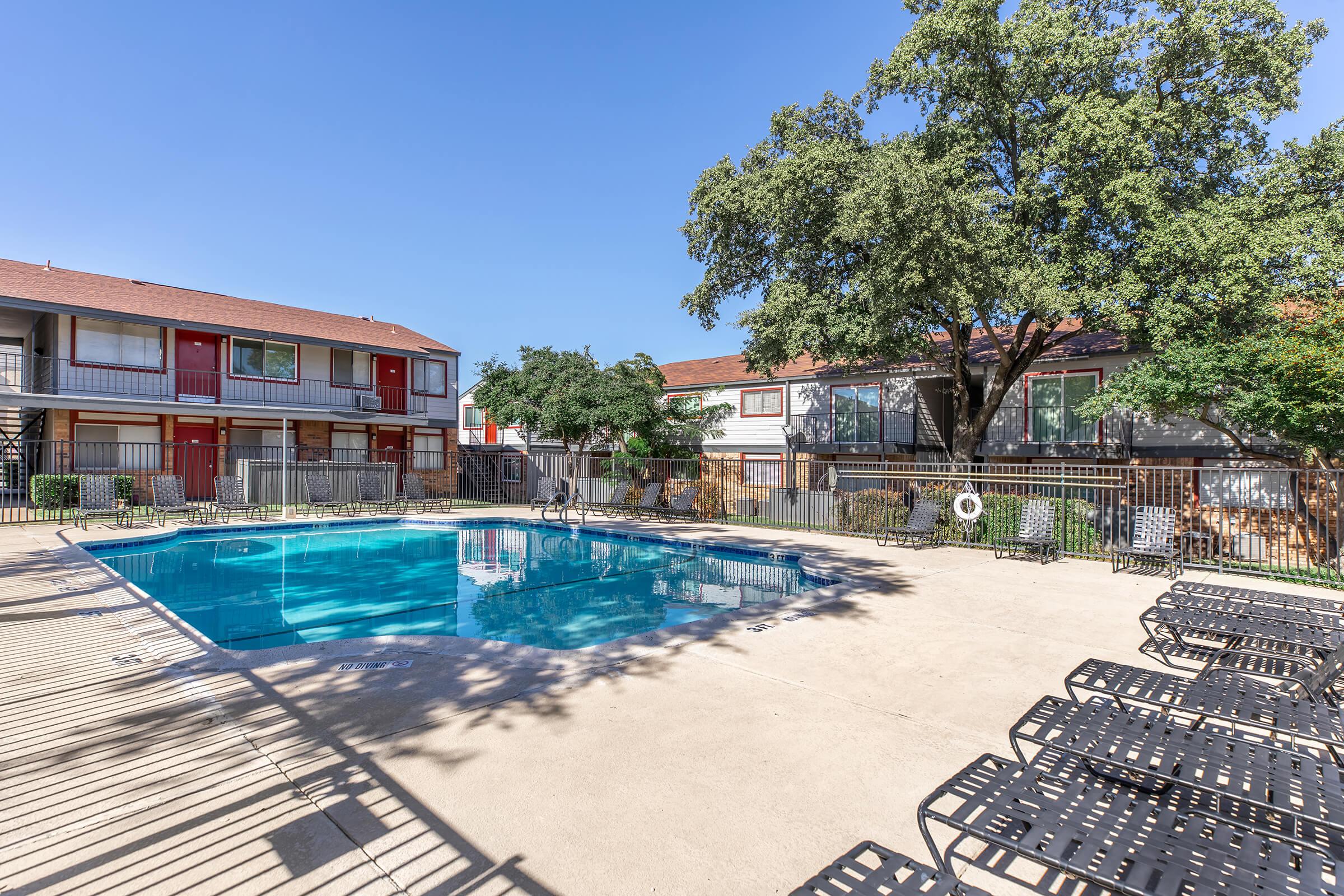 A clear blue swimming pool surrounded by lounge chairs, nestled in a courtyard with two-story buildings. Lush green trees provide shade, and a fence frames the pool area. Bright sunlight illuminates the scene, creating a welcoming outdoor atmosphere.