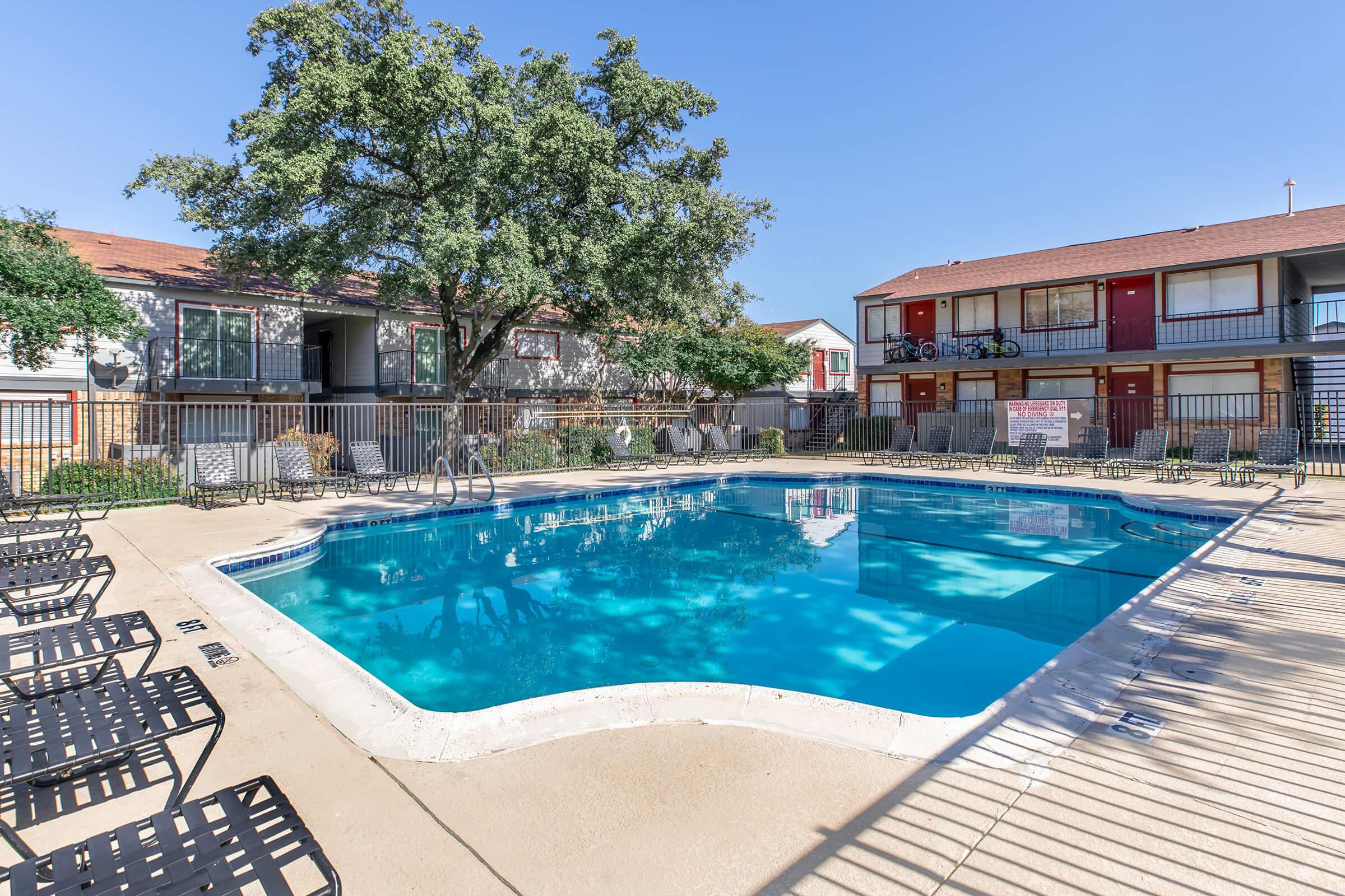 A clear swimming pool surrounded by lounge chairs, with two-story apartment buildings in the background. A large tree provides shade on one side of the pool area. The sky is clear, indicating a sunny day. A fence encloses the pool area, adding to the relaxing atmosphere.
