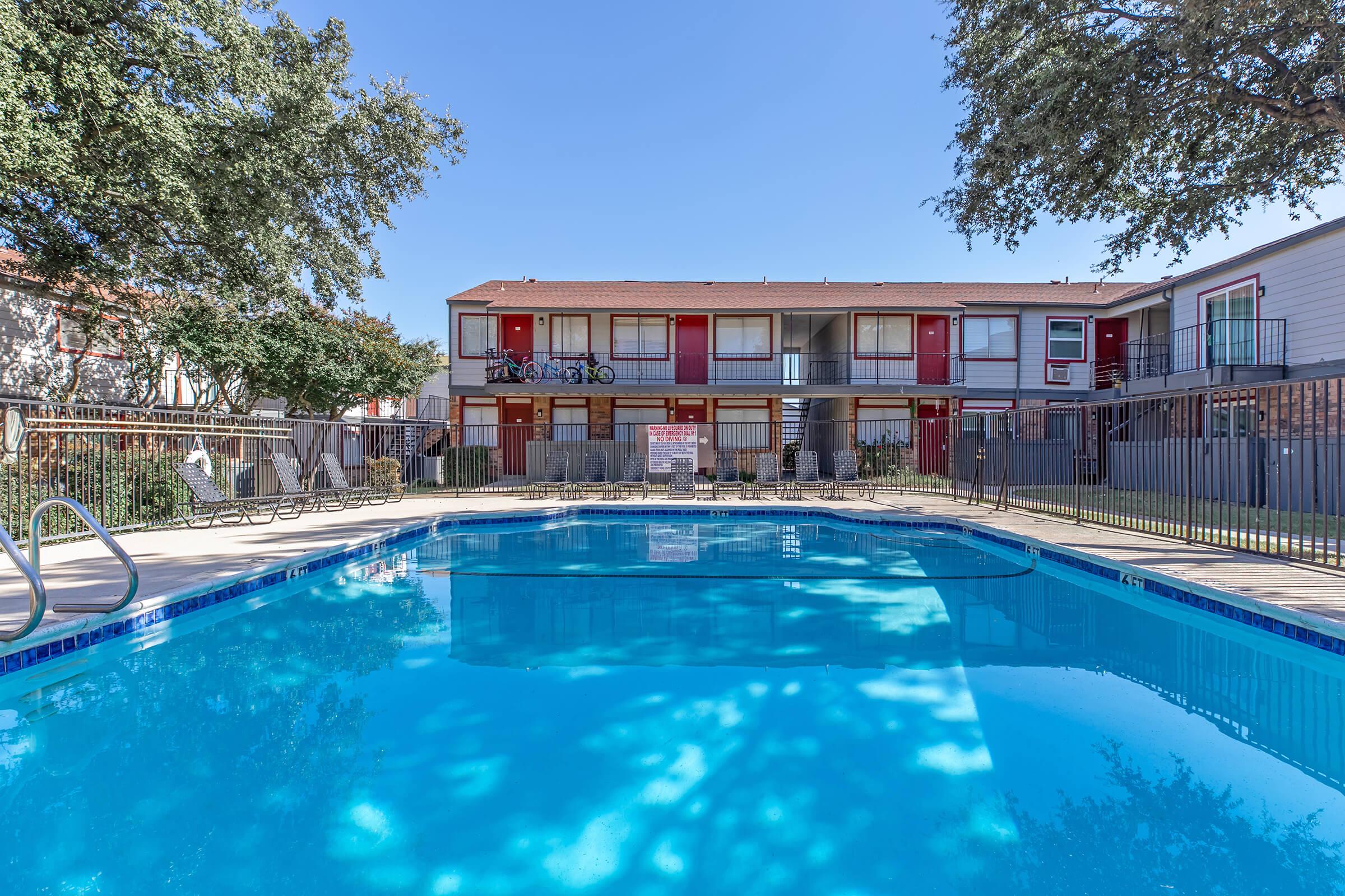 A clear blue swimming pool surrounded by a fenced area with lounge chairs. In the background, a two-story apartment building features red doors and shaded trees. The scene is set on a bright, sunny day with a clear sky.