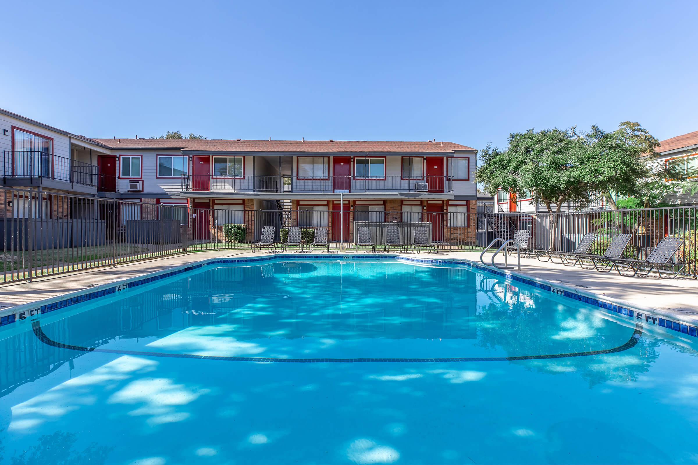 A swimming pool surrounded by lounge chairs and a fence, with an apartment complex in the background. The sky is clear and blue, and trees are visible near the pool area, creating a relaxing atmosphere.