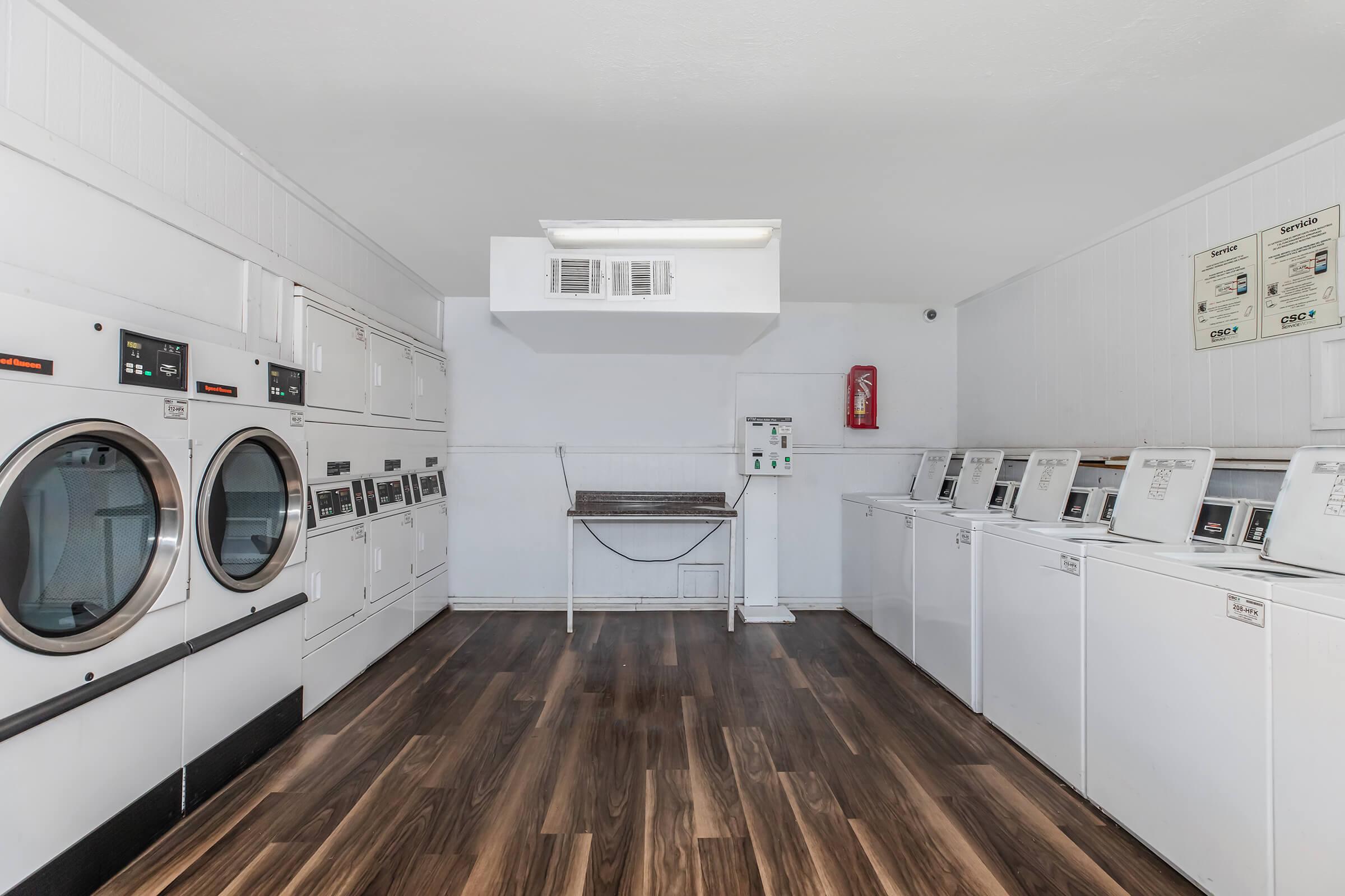 A clean, well-lit laundry room featuring multiple white washing machines and dryers along the walls. There is a folding table in the center and a fire extinguisher mounted on the wall. The flooring is made of dark wood, providing a modern and organized atmosphere for laundry services.