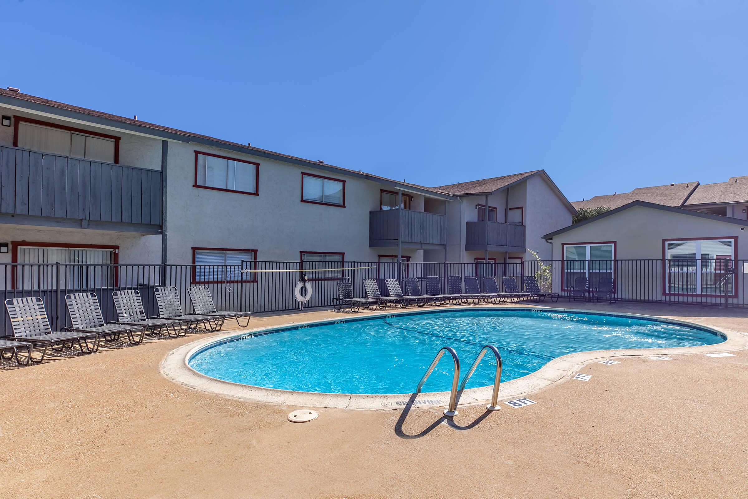 A view of a sunny outdoor swimming pool surrounded by lounge chairs, with a multi-story apartment building in the background. The pool area features a safety buoy and a fenced perimeter, providing a relaxing atmosphere for residents and guests.