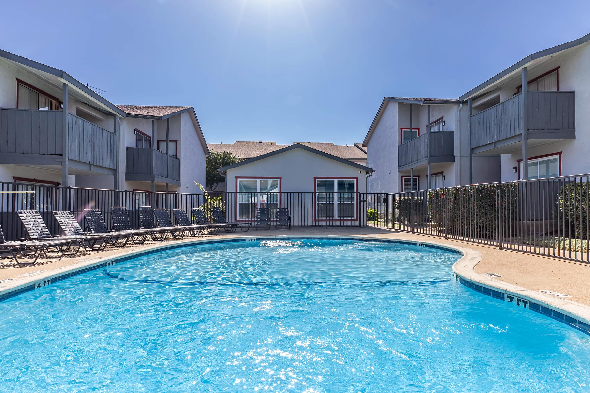 A clear swimming pool surrounded by lounge chairs, with a fenced area. Two buildings with balconies are visible in the background, under a bright blue sky. The scene portrays a relaxing outdoor space.