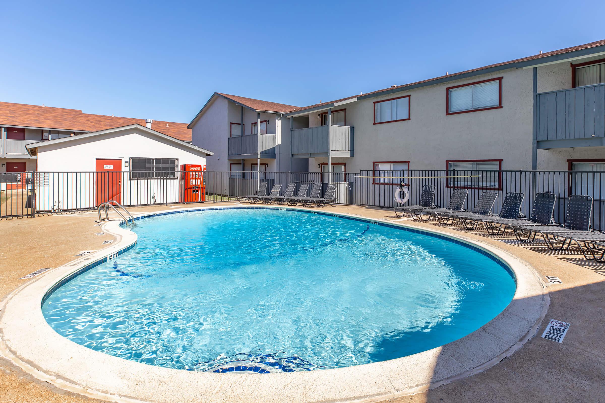 A clear blue swimming pool surrounded by a beige concrete deck, with several lounge chairs arranged nearby. The pool is located in an outdoor area with two-story gray and red buildings in the background, under a bright blue sky.