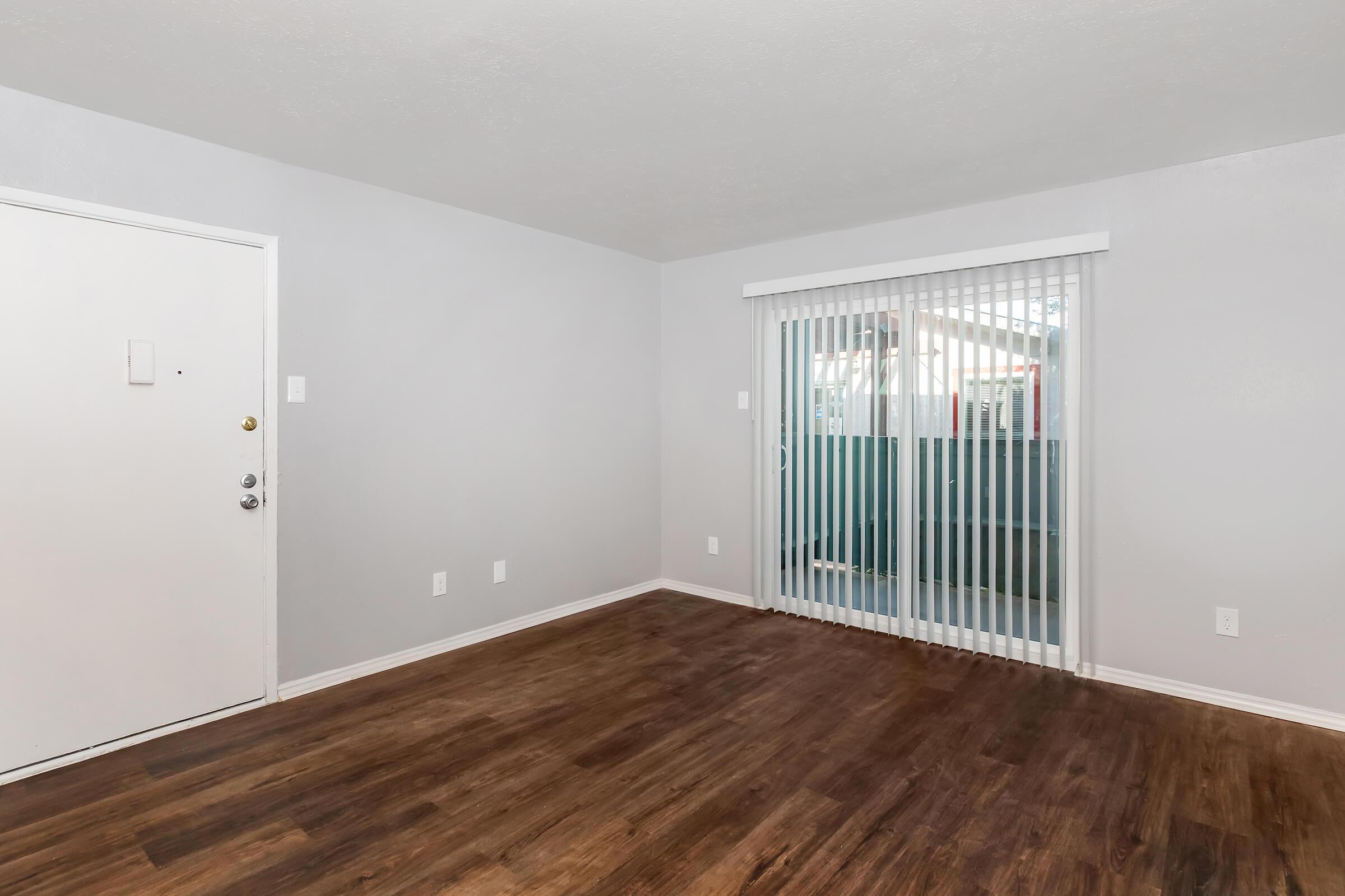 Empty room with light gray walls and laminate flooring. A door is visible on the left, and large sliding glass doors with vertical blinds lead to an outdoor area. The space is well-lit and features minimal furnishings, creating an open and clean atmosphere.