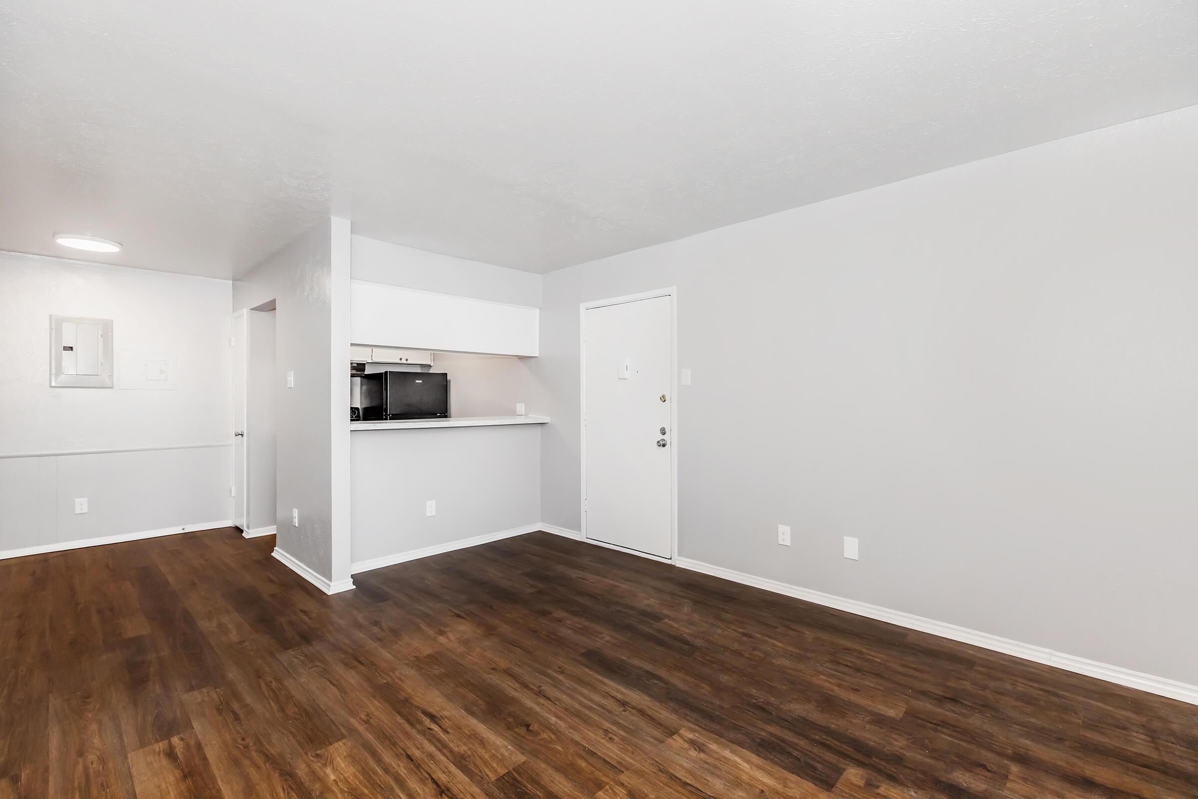 A spacious, modern living area with light gray walls and dark wood flooring. There is a white kitchen area visible on the left, featuring a black microwave. The main entrance door is on the right, and there's a small window above the kitchen counter, providing natural light. The room is unfurnished.