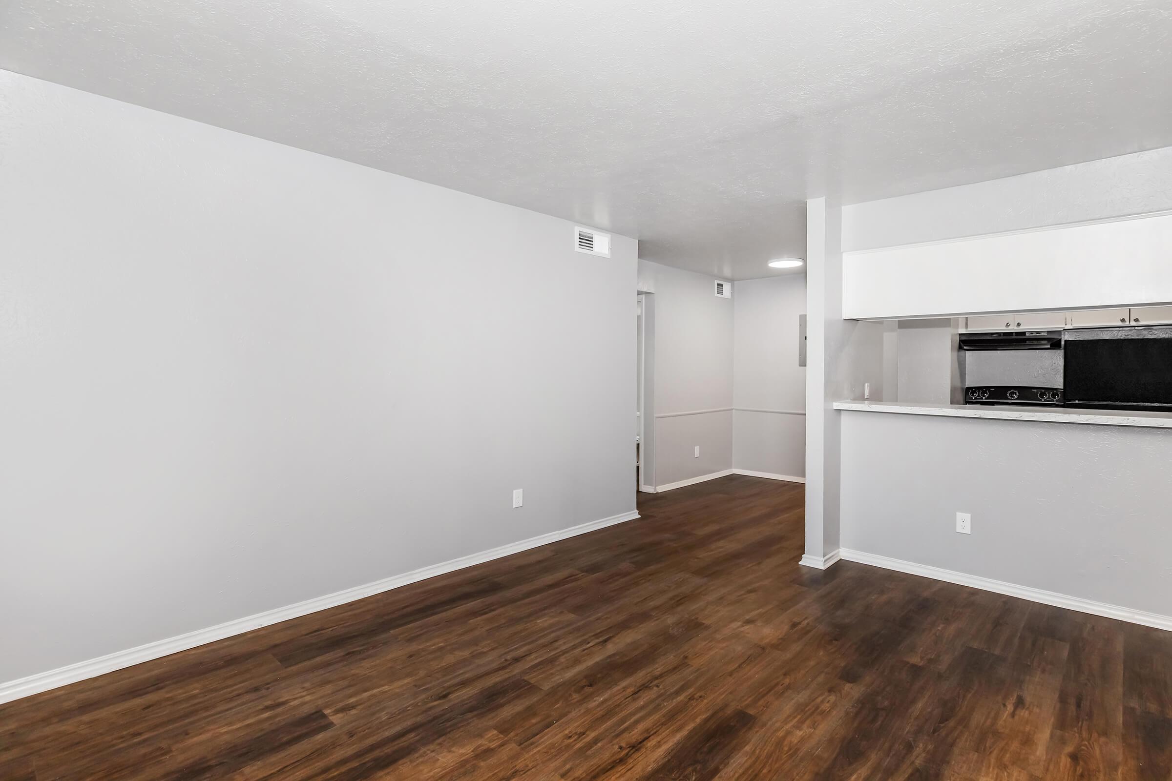 Spacious room with light gray walls and dark wood flooring, featuring an open layout. There is a doorway leading to another room, and a kitchen area with white cabinetry and appliances partially visible in the background. Natural light enhances the bright atmosphere.