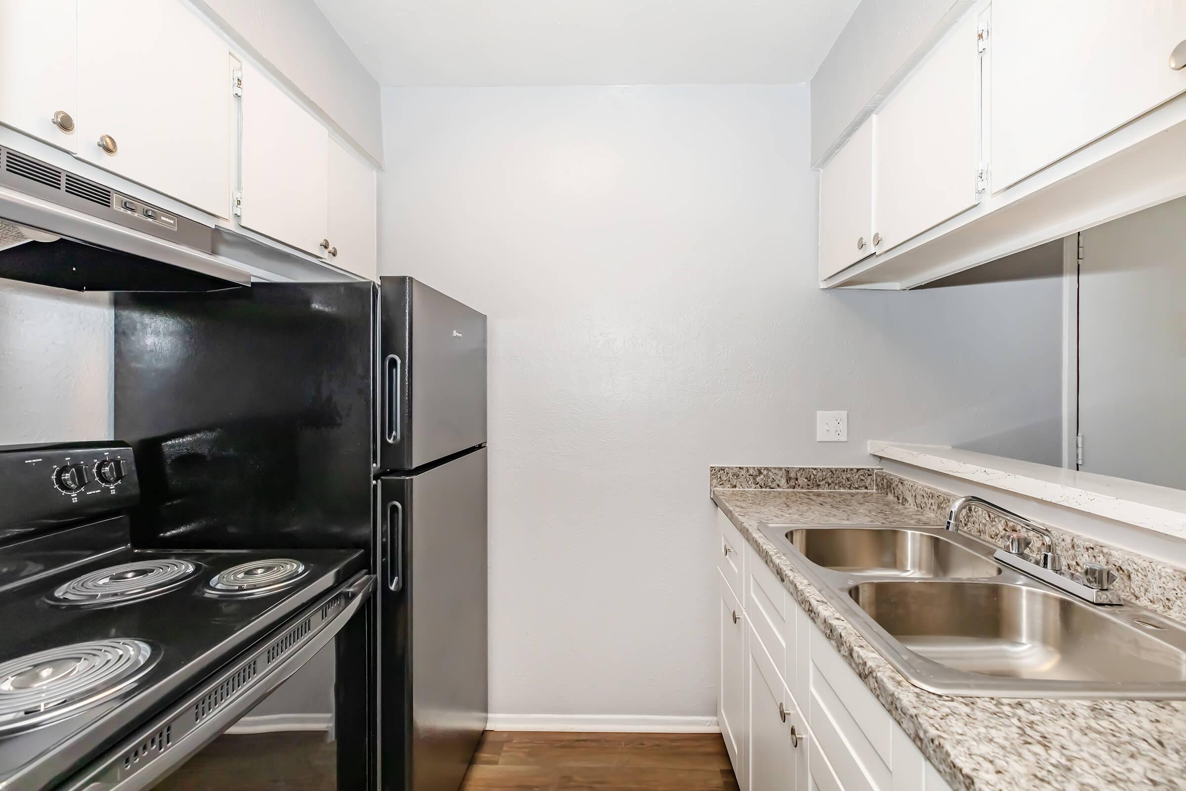 A modern kitchen featuring white cabinets, a black refrigerator, a black stove with an oven, and a double sink. The countertops are made of granite, and the walls are painted light gray. The kitchen is well-lit and has a clean, minimalistic design.