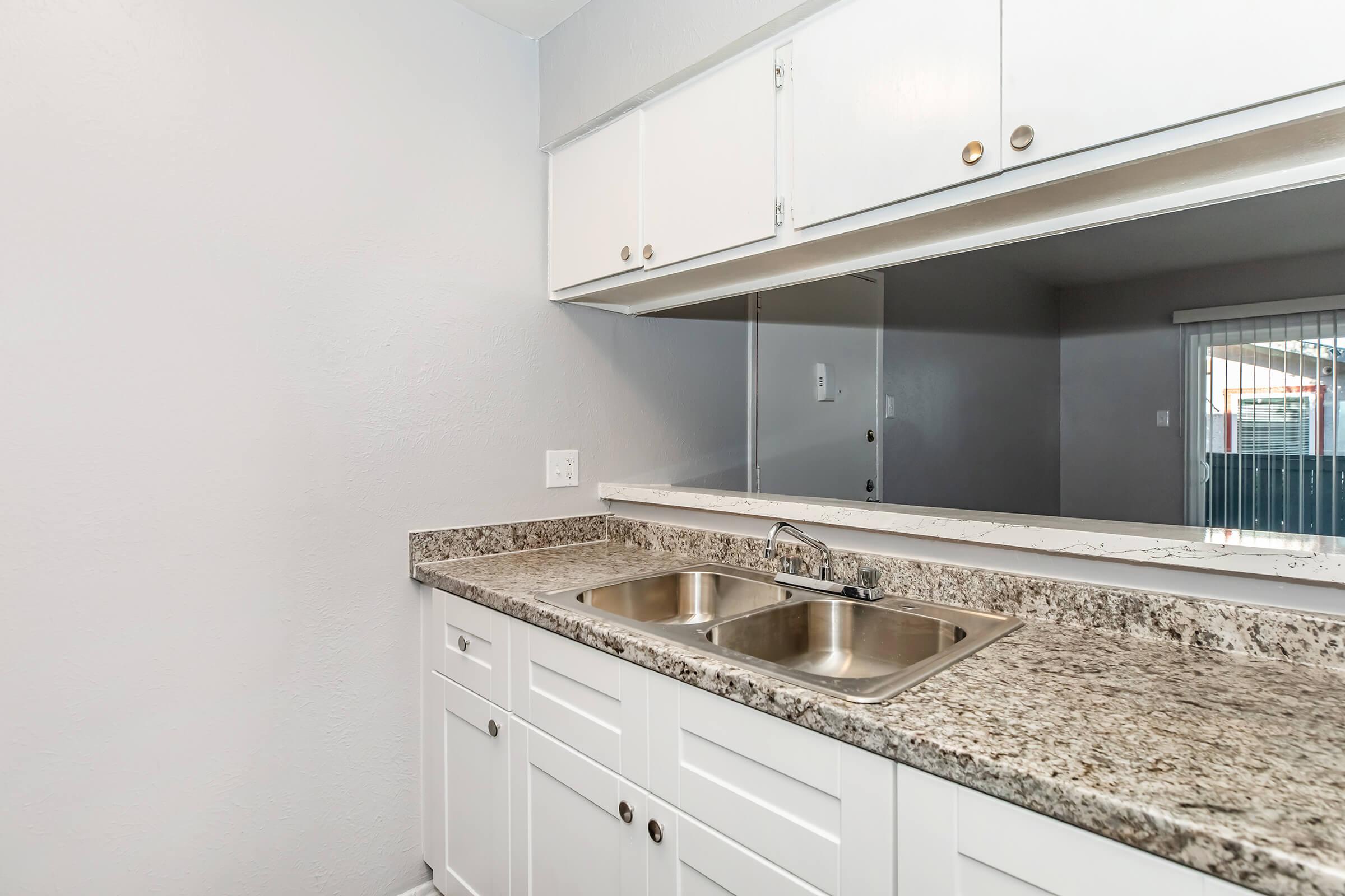 A clean, modern kitchen featuring white cabinetry, a dual sink, and a granite countertop. The wall behind the sink is mirrored, enhancing the sense of space. Soft gray walls provide a neutral backdrop, and natural light comes in from an adjacent area.