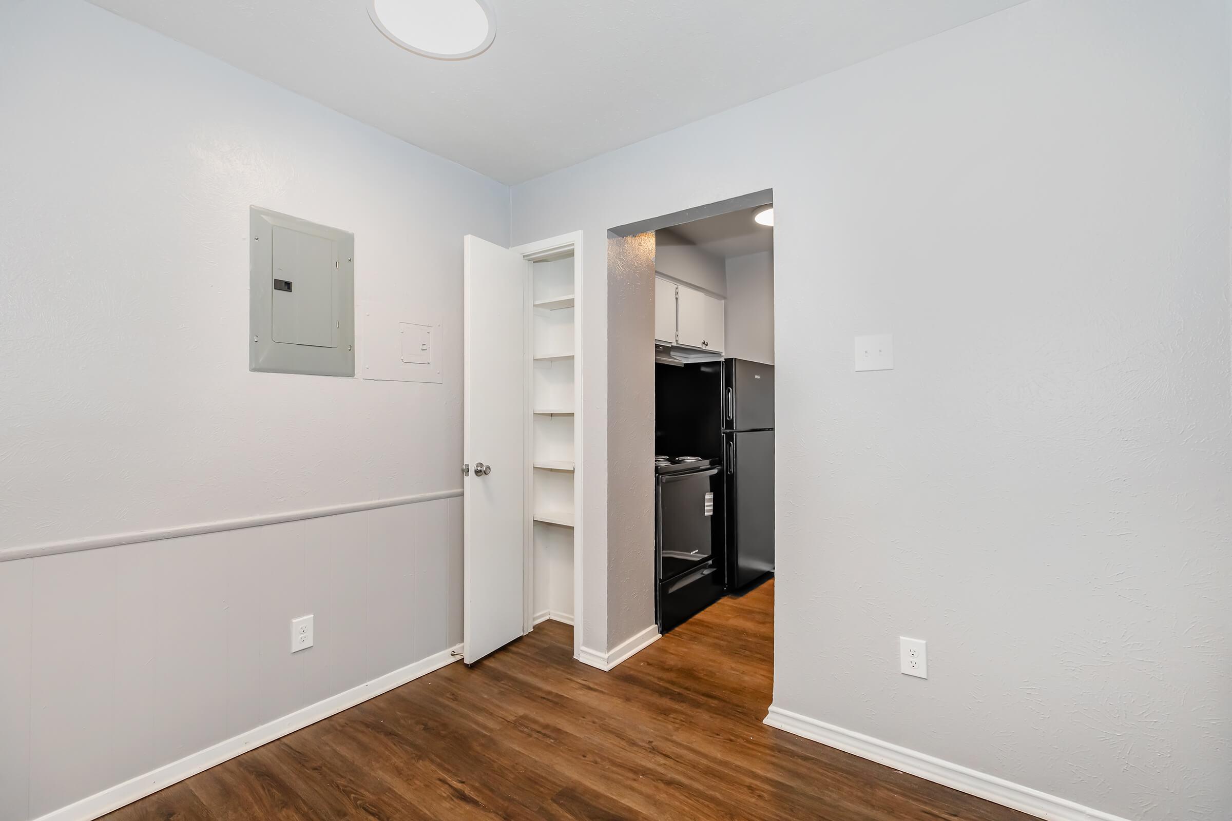 A small, dimly lit room with light gray walls and wood-like flooring. On the left, there is an electrical panel and a closet. To the right, there is an open doorway leading to a kitchenette that features a black refrigerator. The space is tidy and minimalistic.