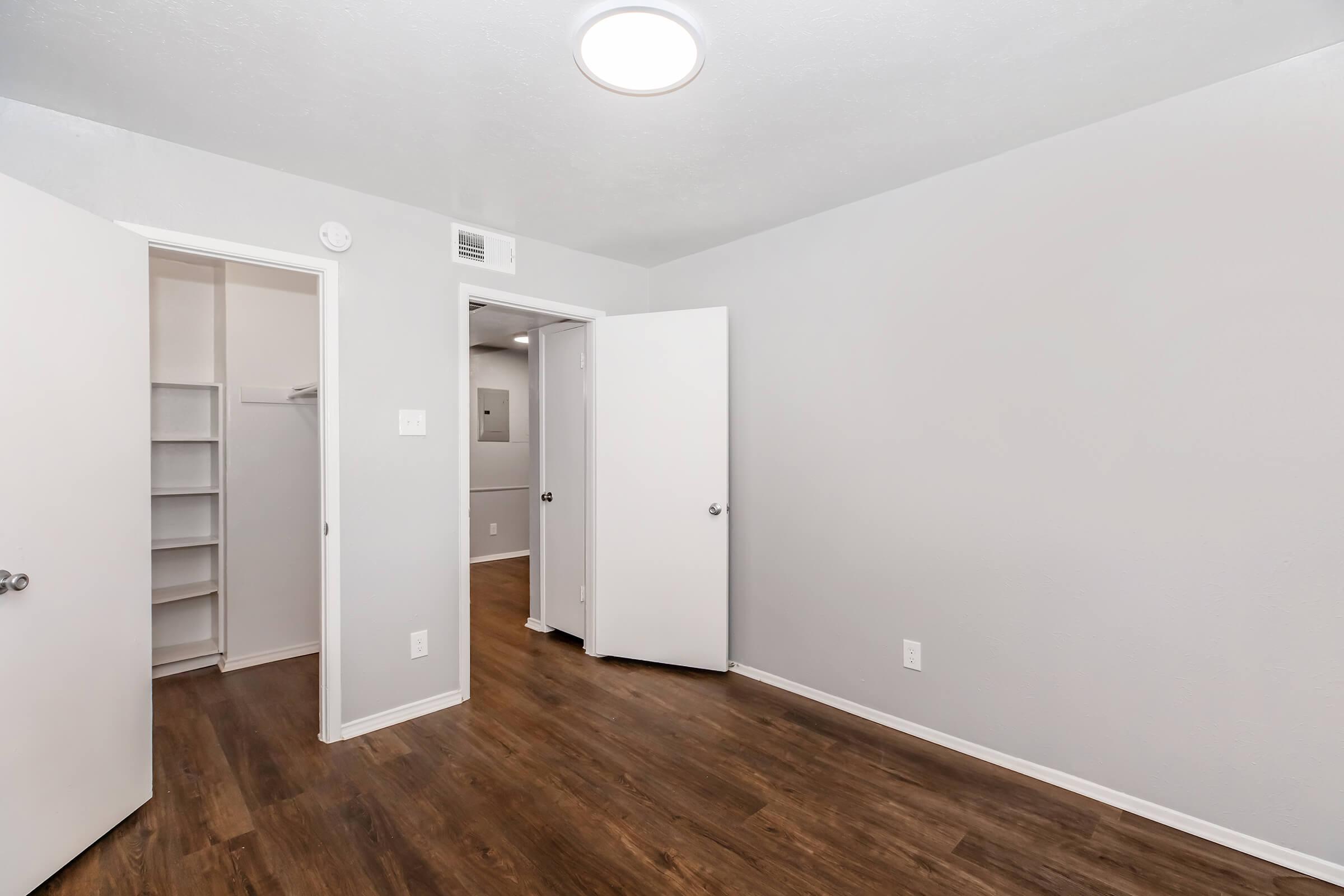 A view of a spacious, empty room with gray walls and a wooden floor. There are two doors: one leading to a closet with shelving and the other to an adjoining area. The ceiling features a circular light fixture, and the overall space appears bright and well-maintained.