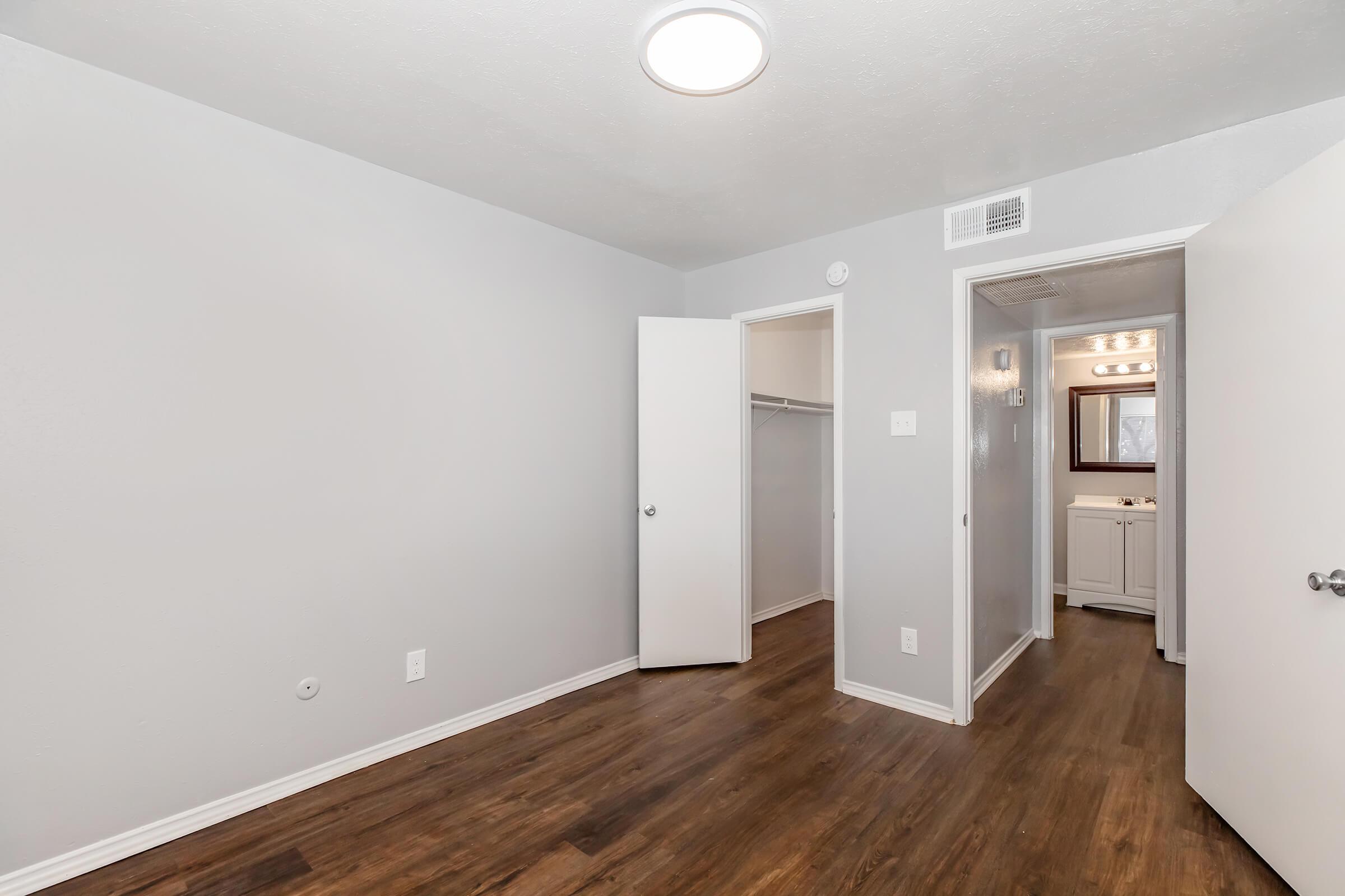 A well-lit, empty room with gray walls and brown laminate flooring. One door is open to a closet, while another leads to a bathroom area with bright lighting visible in the distance. The overall space appears clean and recently updated.