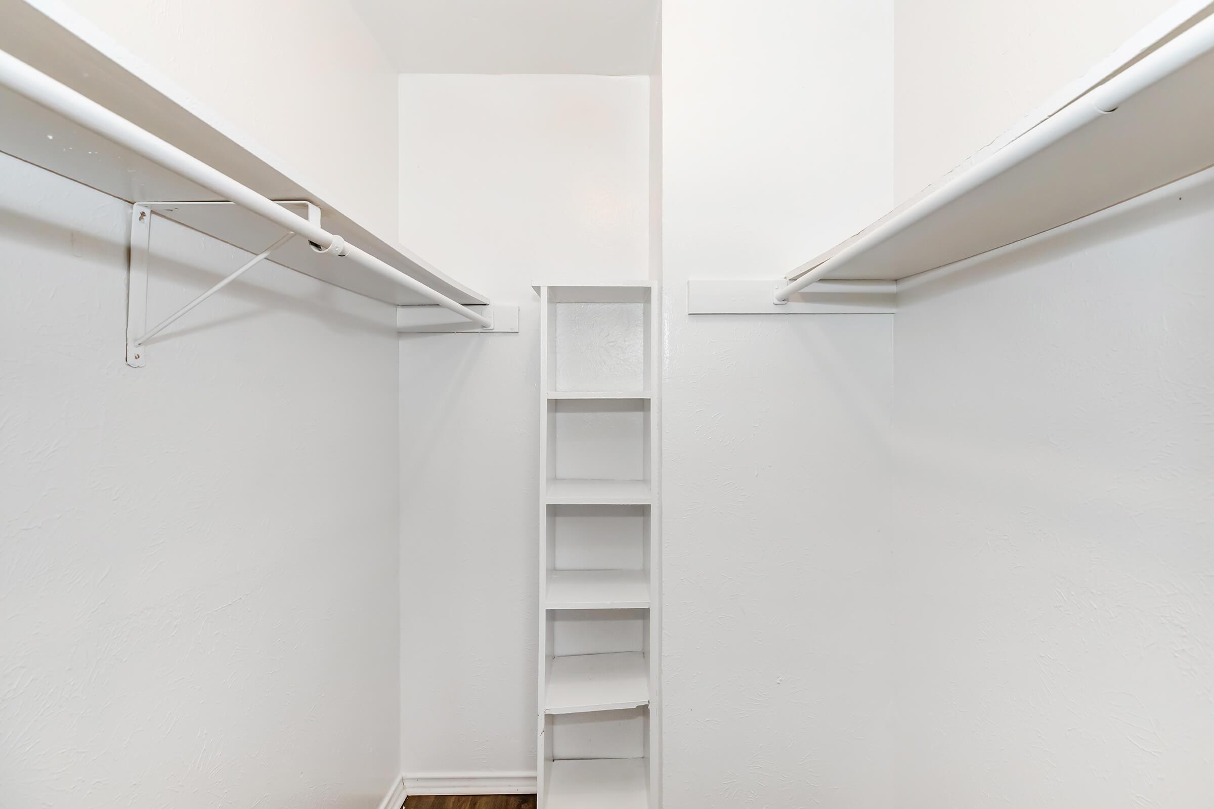 An empty closet featuring white walls and two hanging rods on either side, with a small shelf unit against the back wall. The floor is wooden, and the overall space appears clean and unobstructed.