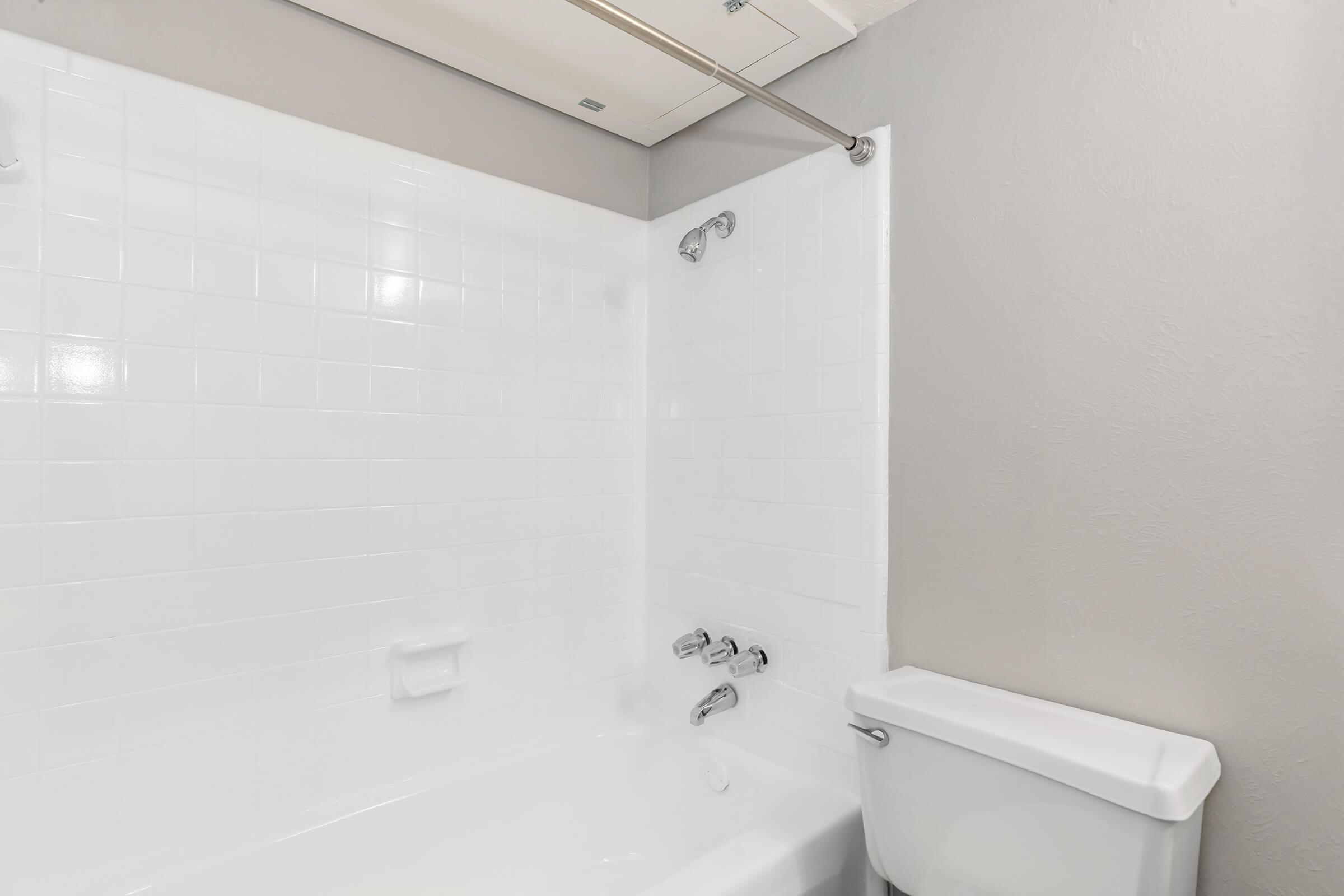 A clean, minimalistic bathroom featuring a white tiled shower area with a bathtub, a showerhead, and wall-mounted fixtures. A white toilet is visible beside the tub against a light gray wall. The overall design is simple and modern, emphasizing functionality and cleanliness.