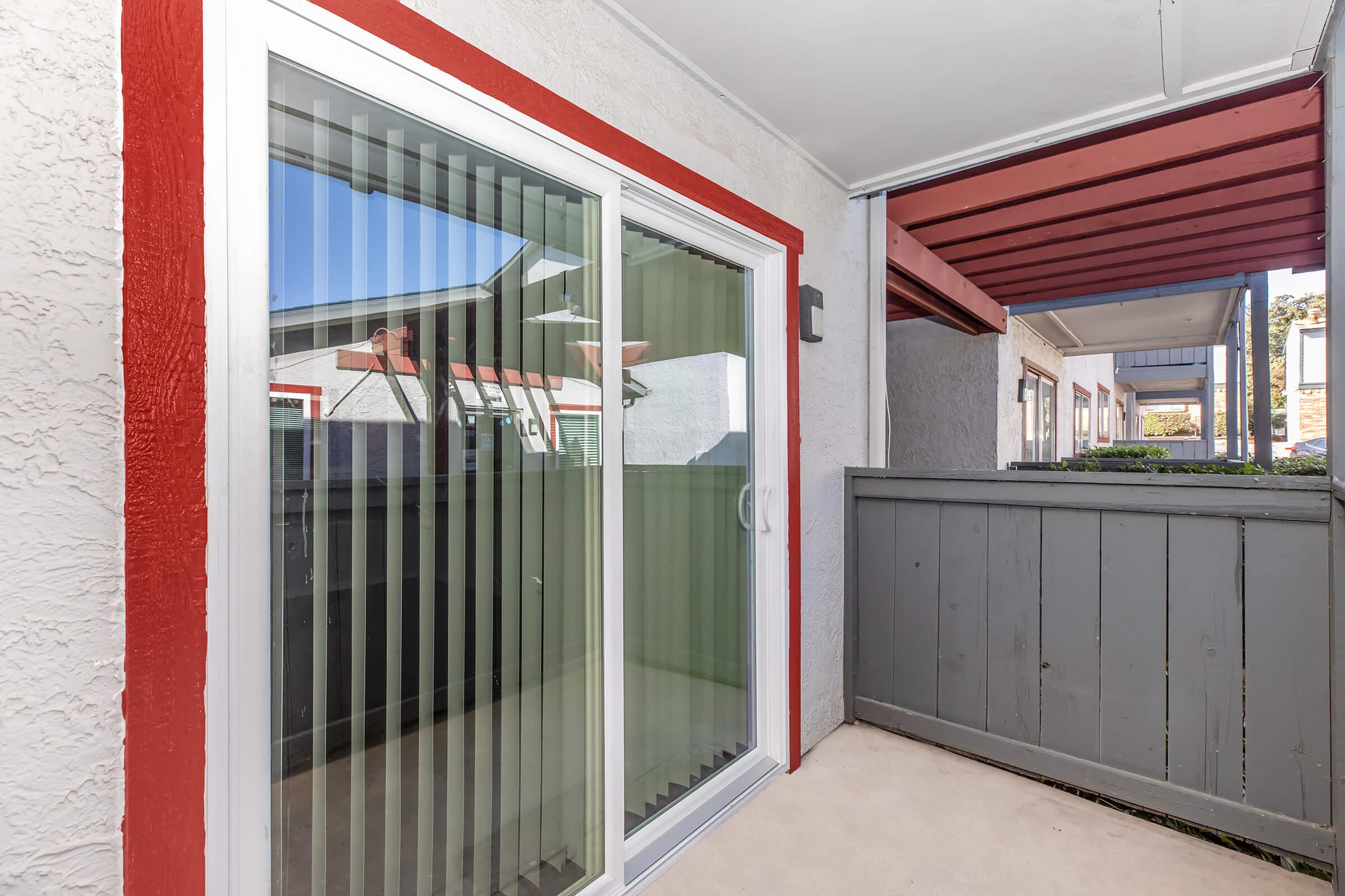 Sliding glass door with vertical blinds, leading to a small patio area. The exterior features light-colored walls with a red trim and a gray wooden fence. Visible buildings in the background suggest a residential complex setting. The scene is bright and well-lit.