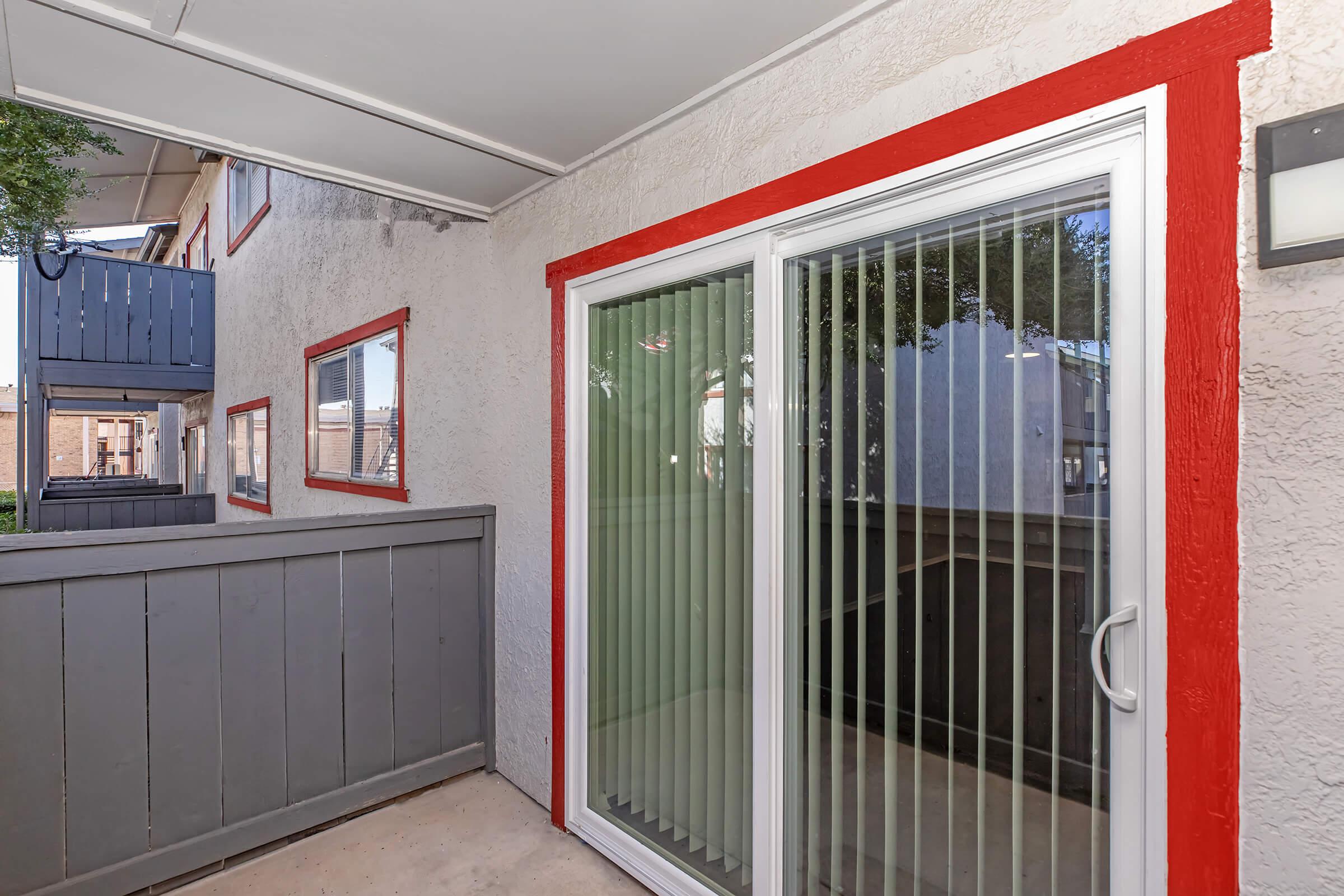 Exterior view of a patio area featuring sliding glass doors with vertical blinds. The walls are painted light gray with red trim, and a wooden fence is present. In the background, there are other apartments visible, creating a residential setting. The area is well-lit and inviting.