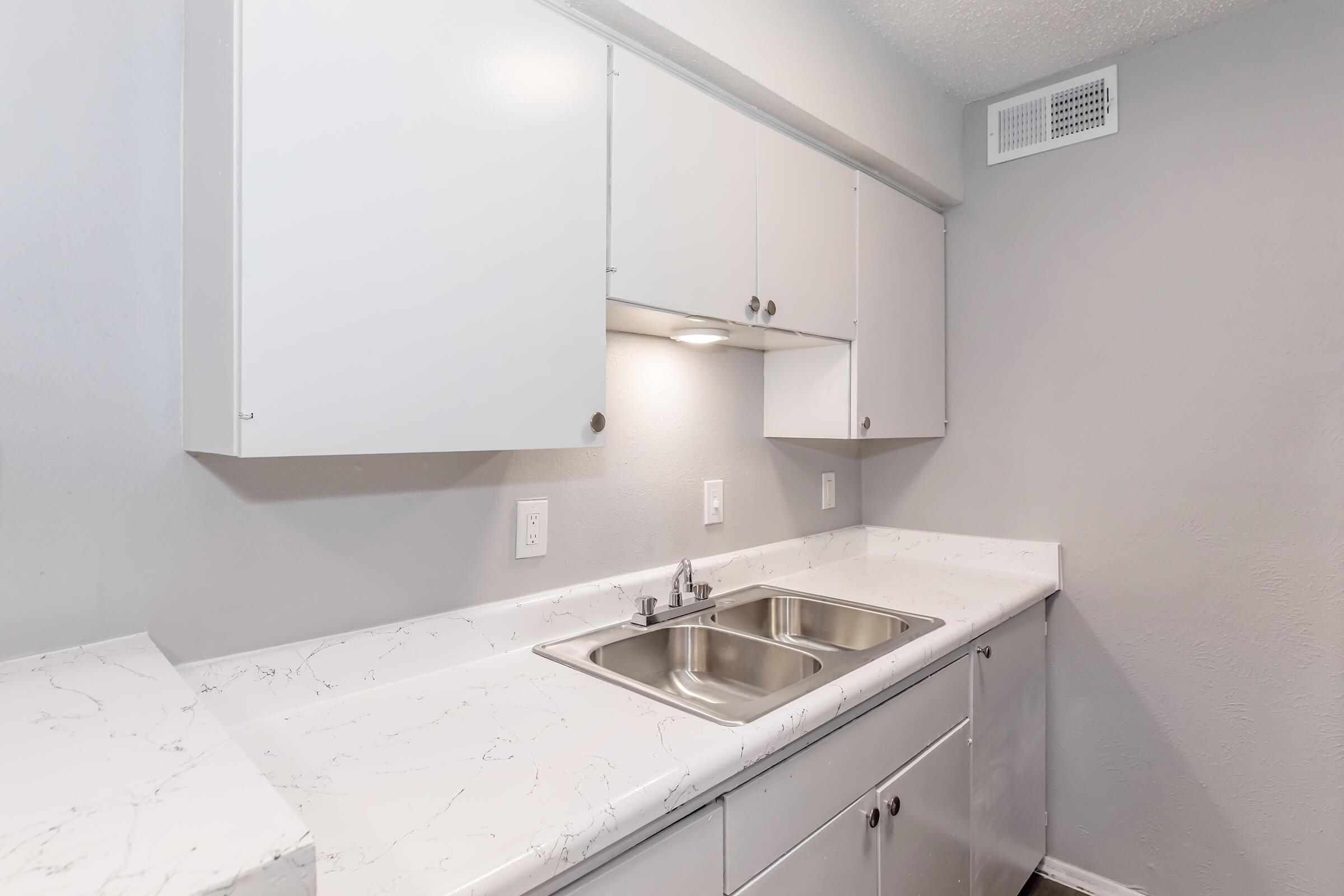 A modern kitchen featuring white cabinetry and a marble-patterned countertop. There are two stainless steel sinks with a faucet, under-cabinet lighting, and a neutral gray wall. The overall design is sleek and minimalistic, creating a clean and inviting space.