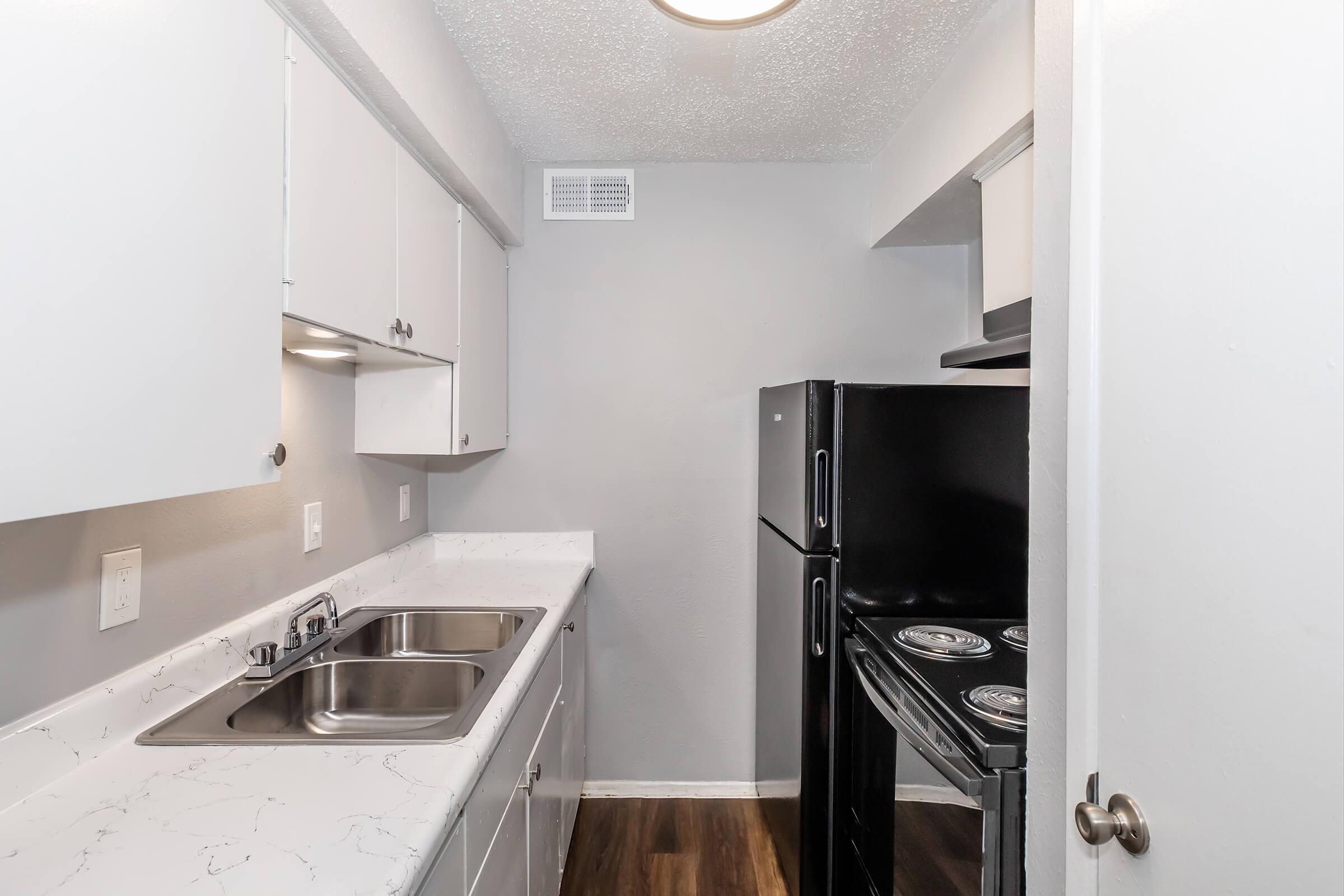 A modern kitchen featuring white cabinets, a double stainless steel sink, and black appliances, including a refrigerator and stove. The wall and countertop have a light color scheme, creating a clean and bright atmosphere. The flooring is dark wood, adding contrast to the overall design.