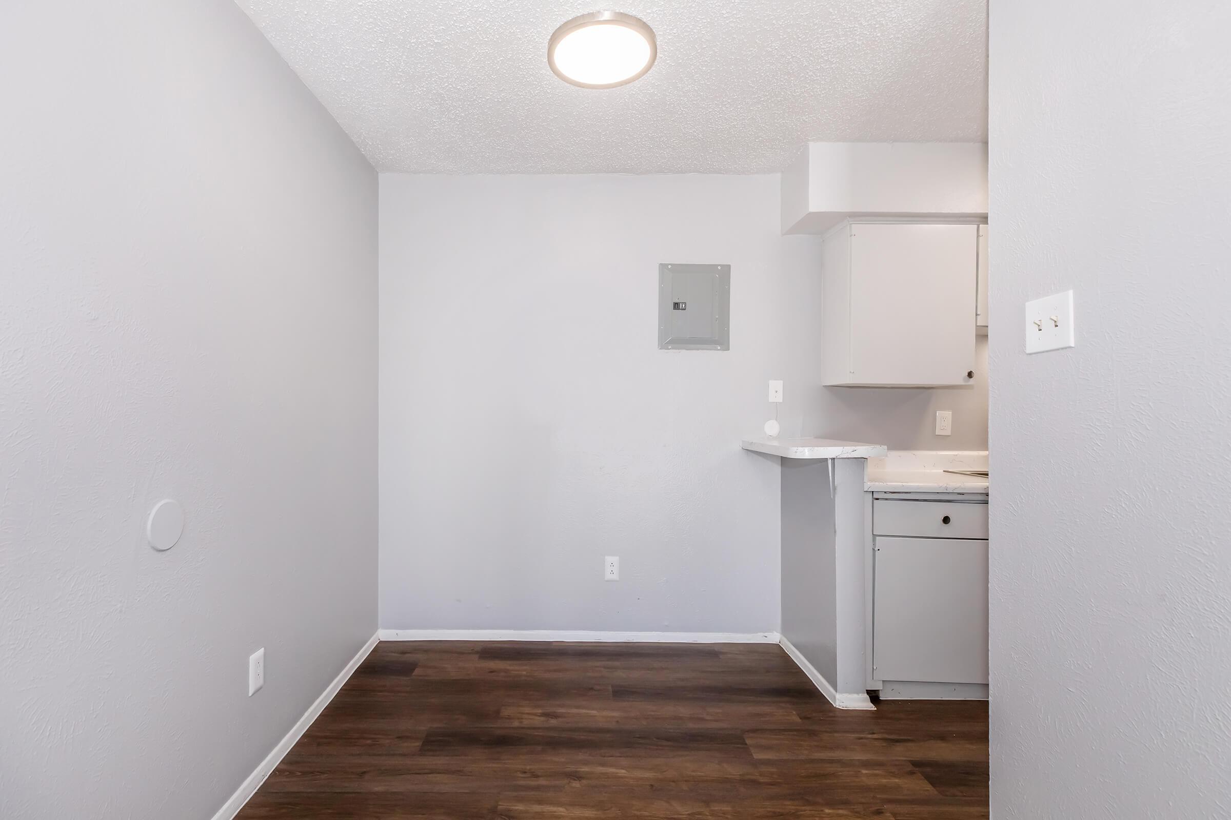 A small, empty room with light gray walls and dark wooden flooring. There's a round ceiling light and an open kitchen area with white cabinets and a countertop. A circuit breaker panel is visible on one wall, and there are no furnishings or decorations in the space, giving it a clean, minimalist look.