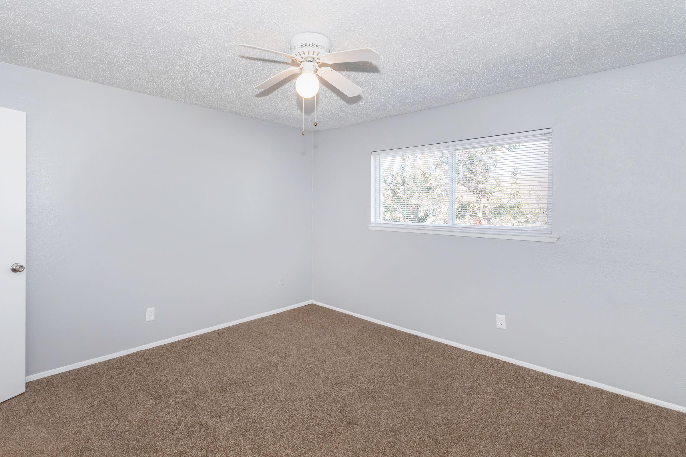 A light gray bedroom featuring a ceiling fan and a window with white blinds. The walls are painted light gray, and the floor is covered in tan carpet. There is a white door visible on the left side of the image, and the overall space is well-lit with natural light coming through the window.