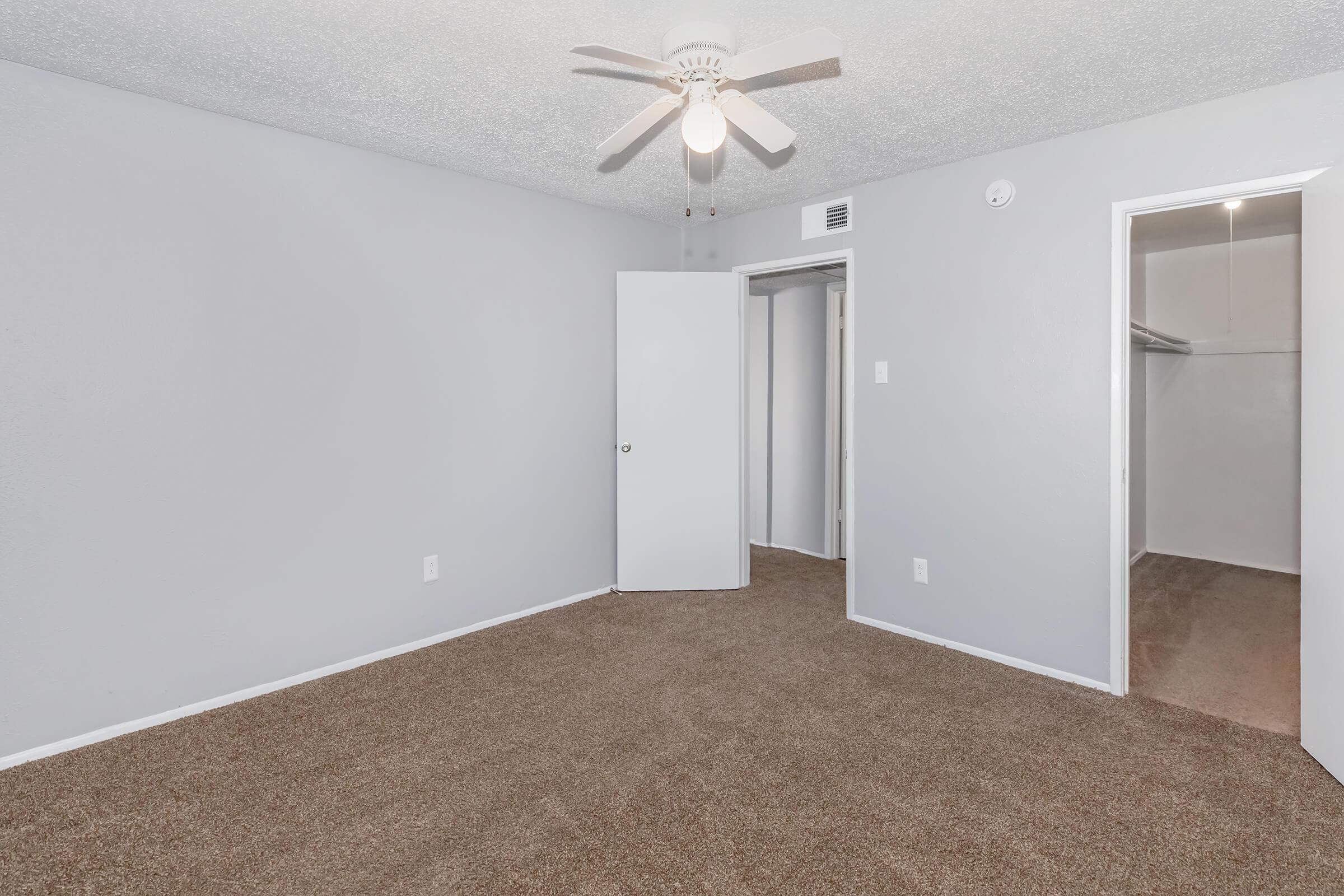 A light gray room with a carpeted floor and a ceiling fan. There is a single door on the left leading to a closet, and another door on the right leading to a separate space. The walls are painted light gray, creating a neutral and spacious atmosphere.