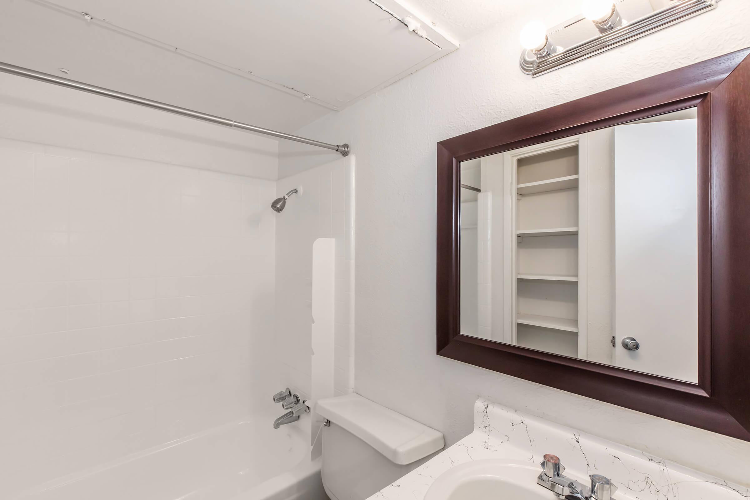 Bright, clean bathroom featuring a white bathtub and shower combo, a modern mirror with a wooden frame, and a marble countertop with a sink. The walls are minimally decorated, and there are built-in shelves visible in the background, creating a spacious and inviting atmosphere.