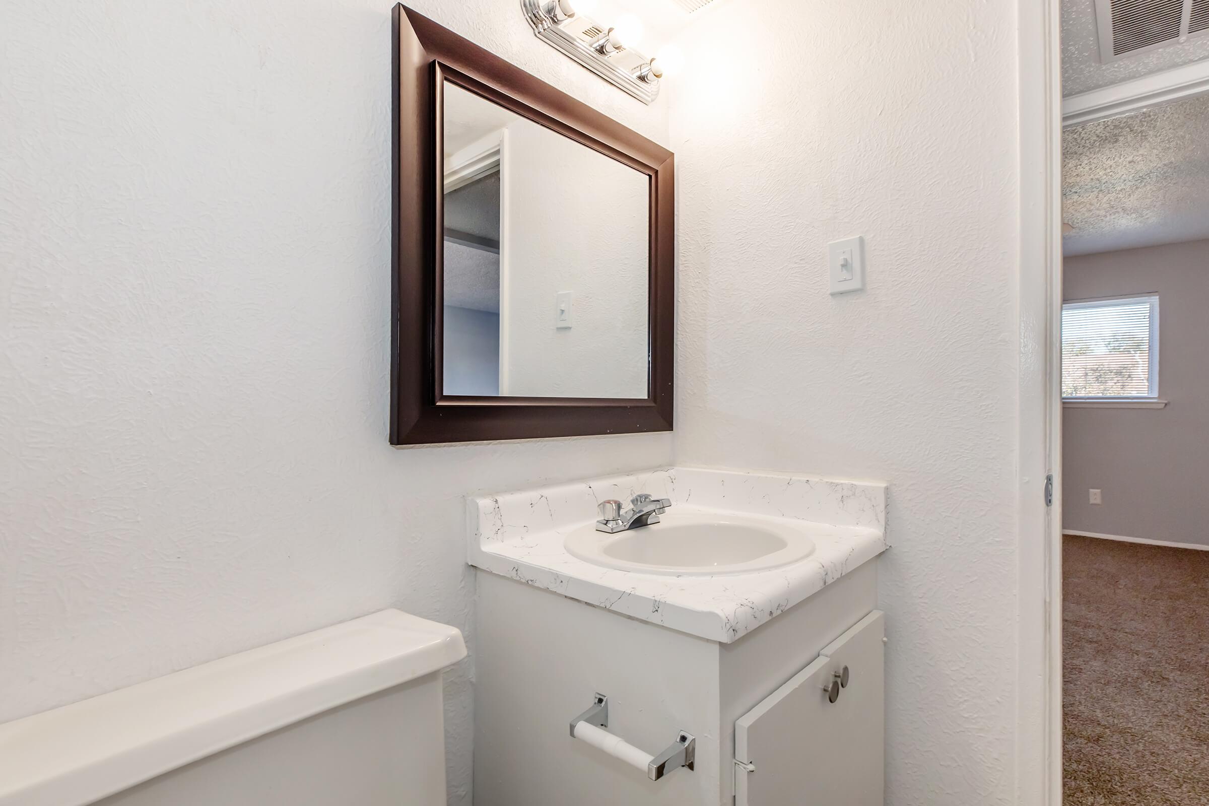 A small bathroom featuring a white vanity with a round sink, a large mirror above, and a light fixture. The wall is painted white, and there is a toilet to the left. The flooring appears to be carpeted in a light brown color, and a window is visible in the background, providing natural light.