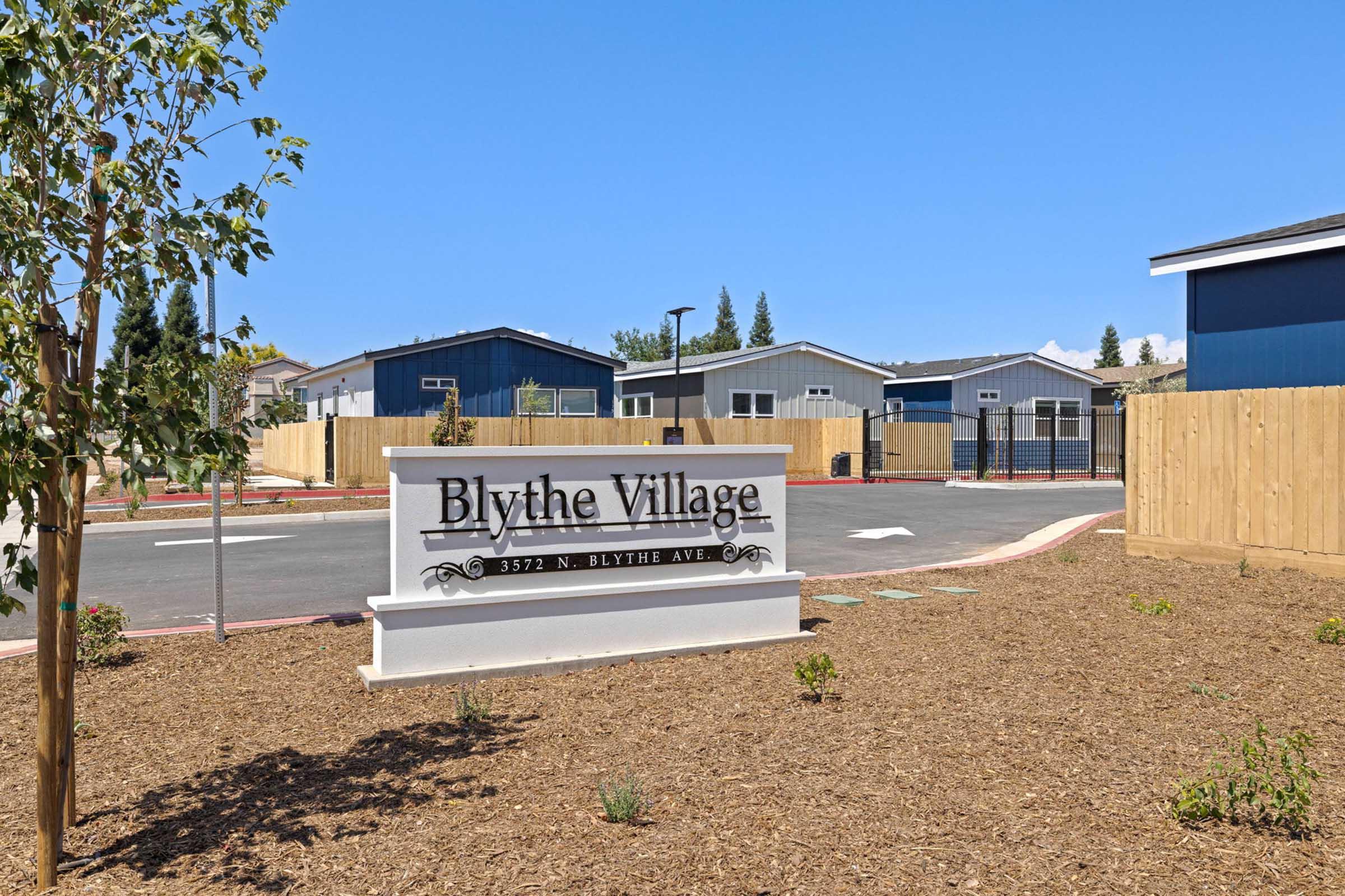 Sign for Blythe Village with landscaped area, featuring nearby residential buildings and a clear blue sky in the background.