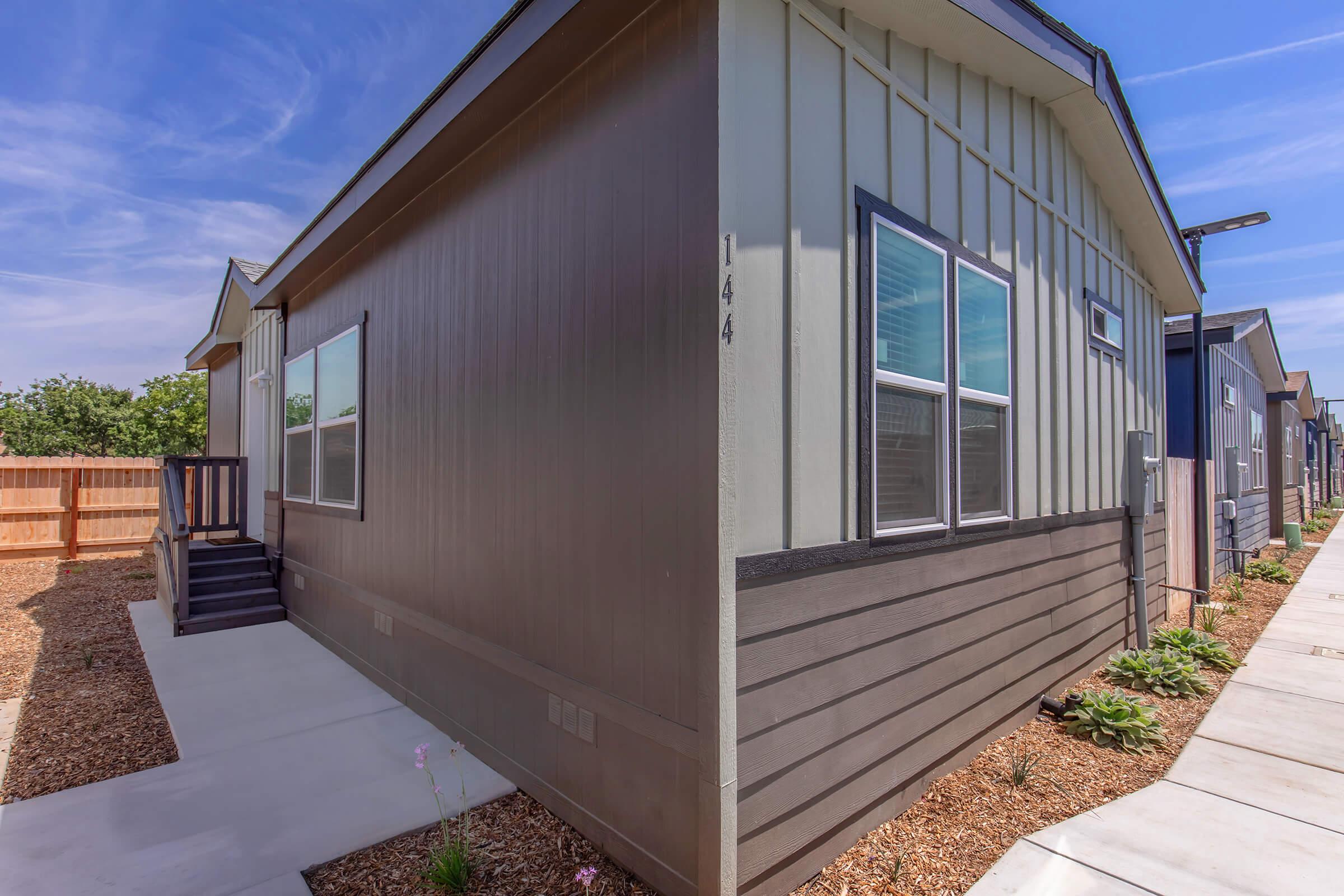 A modern two-story building showcasing a blend of dark and light exterior siding. A small porch with steps leads to a side entrance. The surrounding area features a well-kept pathway and landscaping with small plants, under a clear blue sky. Fencing provides privacy in the background.