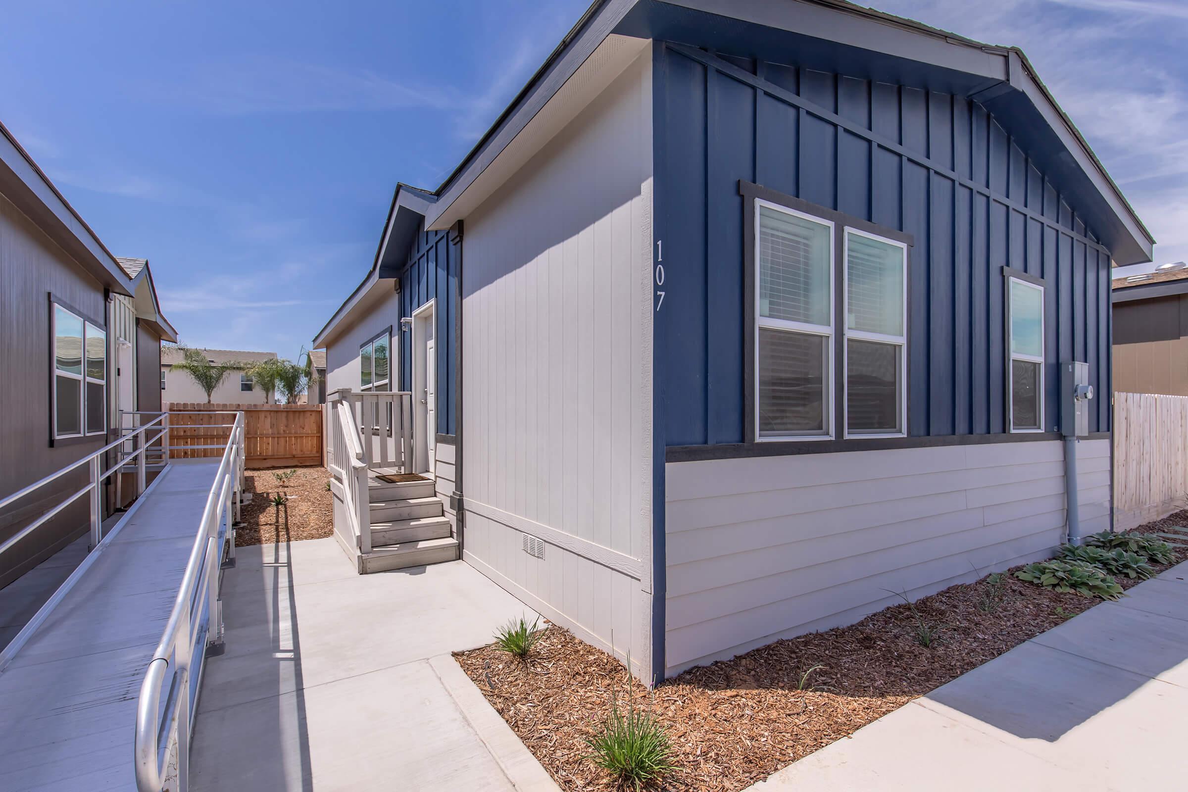 A modern single-story house featuring a blue and gray exterior, with a front ramp for accessibility. It has large windows, a small deck with stairs, and landscaping with small shrubs and decorative gravel paths. The surrounding area includes similar homes and clear blue skies.