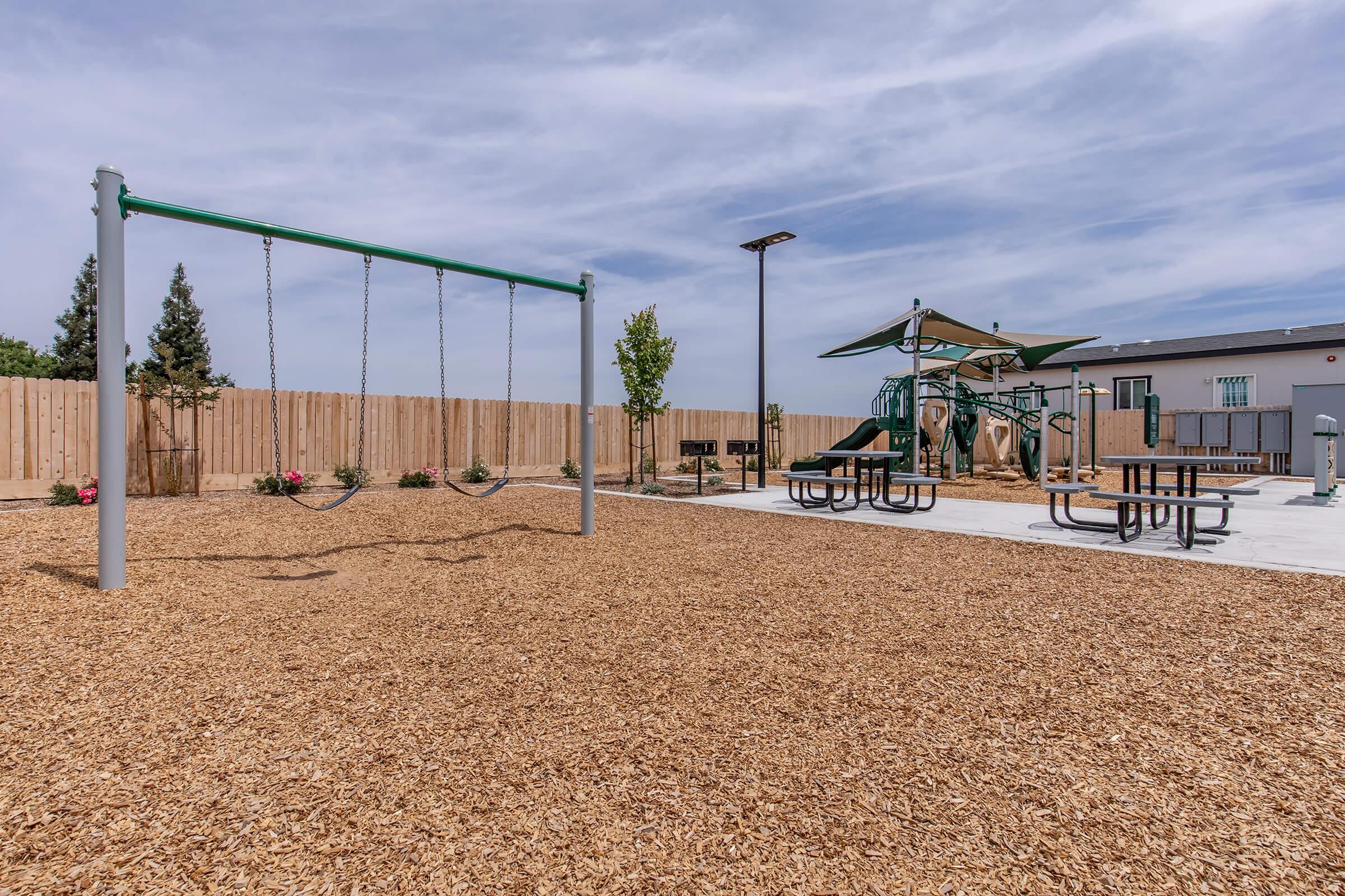 A children's playground featuring swings and a play structure, surrounded by wood chip flooring. There are picnic tables and landscaped areas with small plants and trees against a wooden fence, under a clear sky.