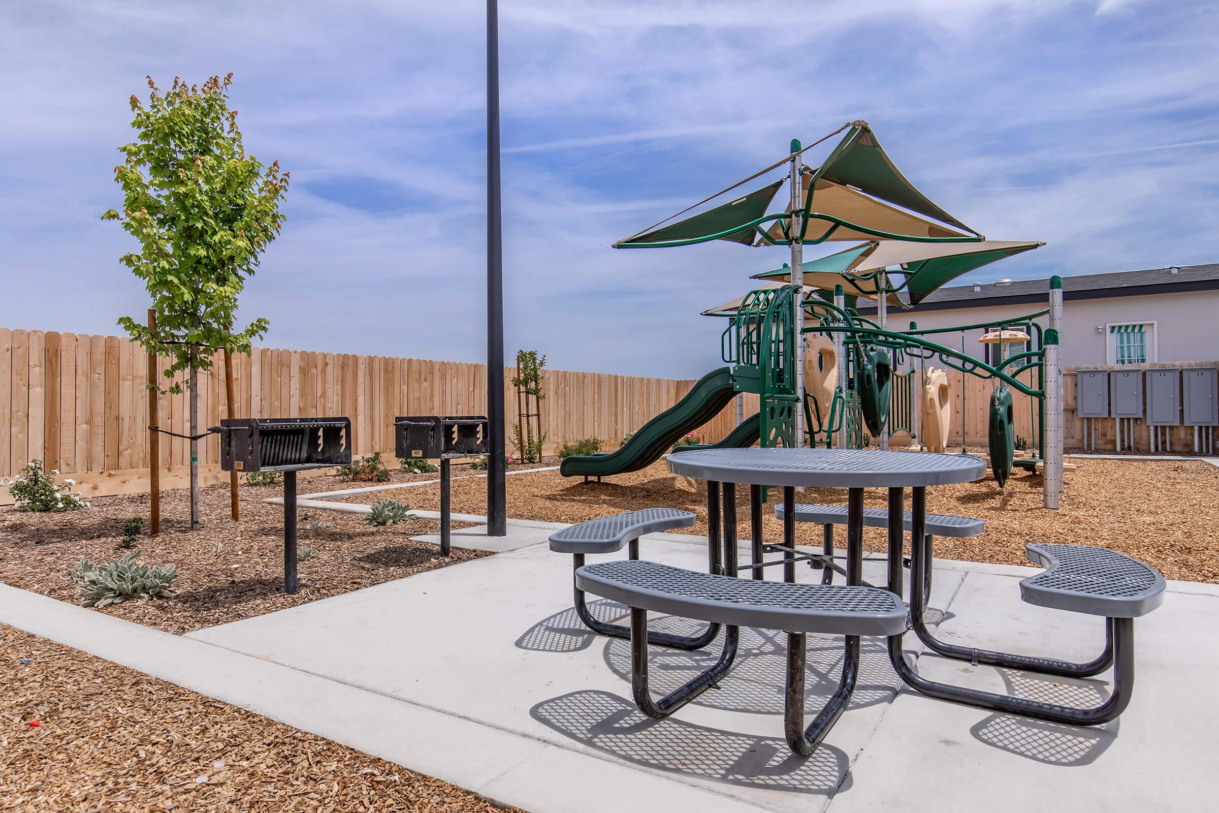 A playground featuring a green play structure with slides, surrounded by a grassy area and garden beds. There are picnic tables and grill stations in a paved area, with a wooden fence in the background and trees adding greenery. The sky is partly cloudy, creating a pleasant outdoor atmosphere.