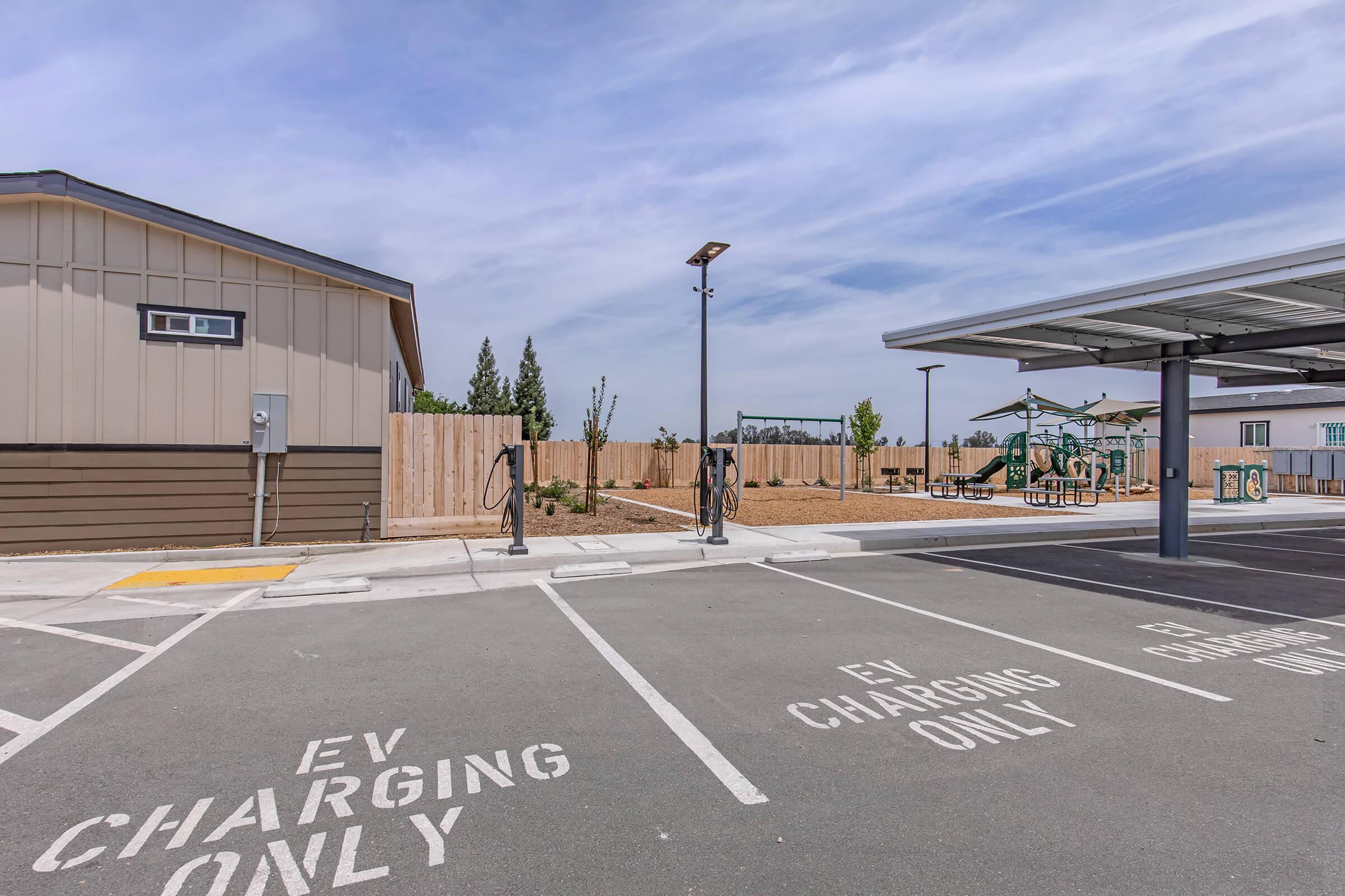 A parking area designated for electric vehicle (EV) charging with clear markings on the pavement. In the background, there's a fenced playground area with playground equipment. The scene includes a building on the left and a shelter for vehicles on the right, under a blue sky with some clouds.