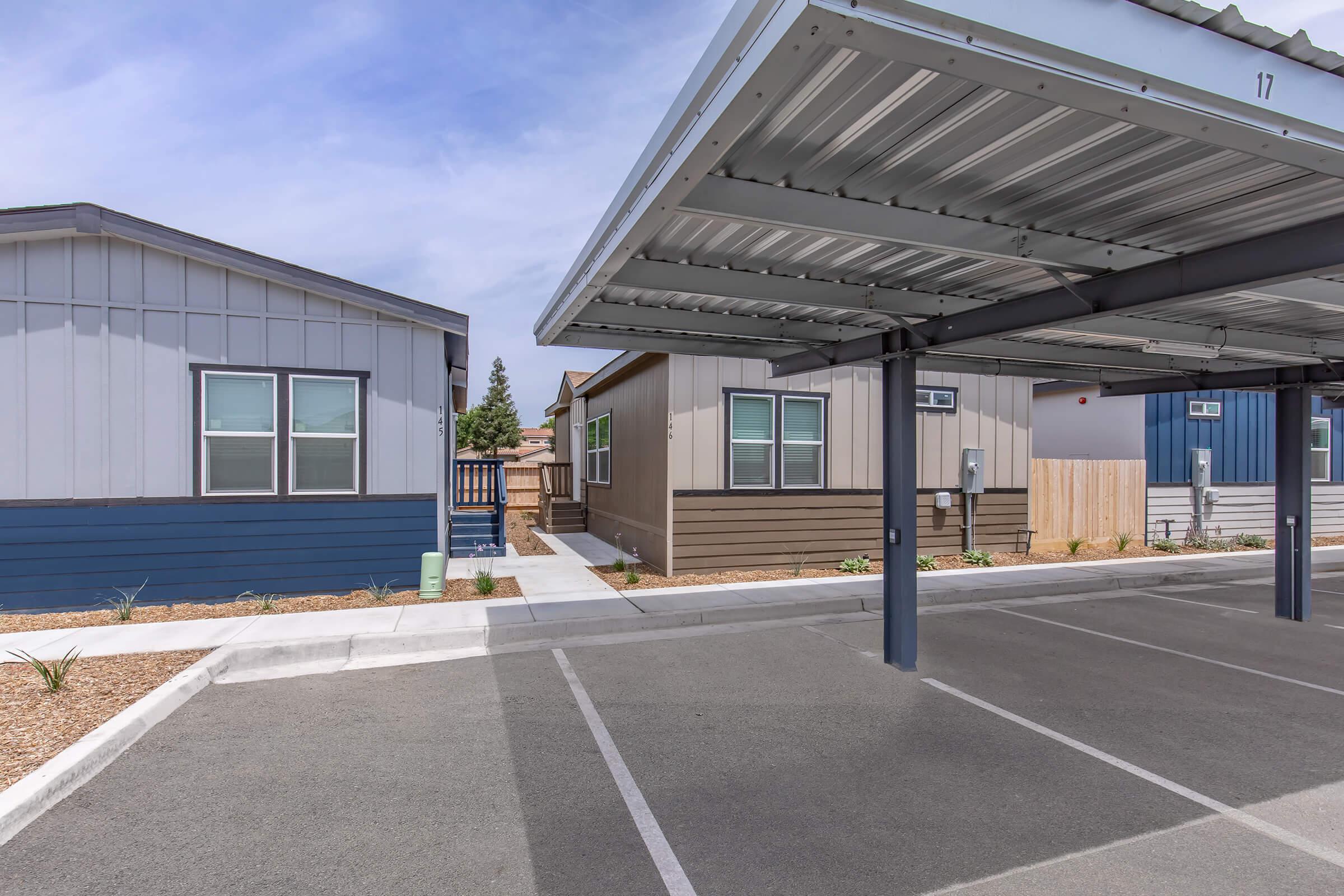 A view of a modern mobile home community featuring two houses in different colors (blue and brown) with a carport. The pathway leading to the homes is lined with plants, and the parking area shows vacant spaces. The sky is partly cloudy, enhancing the serene atmosphere of the neighborhood.