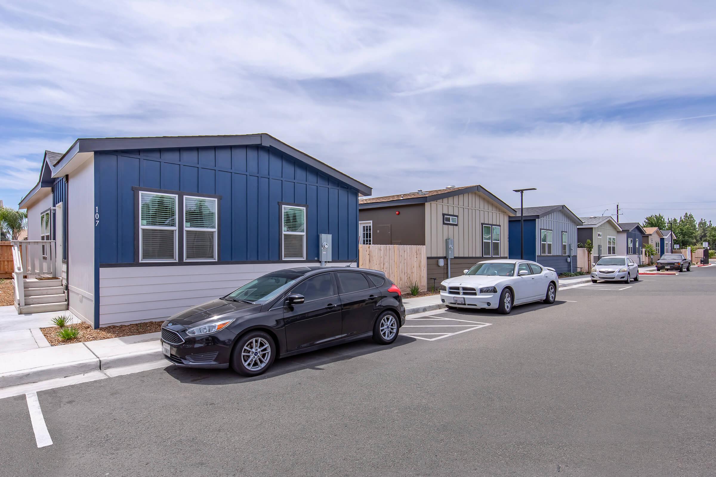 A row of modern manufactured homes along a paved street, with two parked cars in the foreground—one black hatchback and one white sedan. The sky is partly cloudy, and the homes feature a mix of blue and neutral colors. The setting appears to be a suburban neighborhood with landscaped areas.