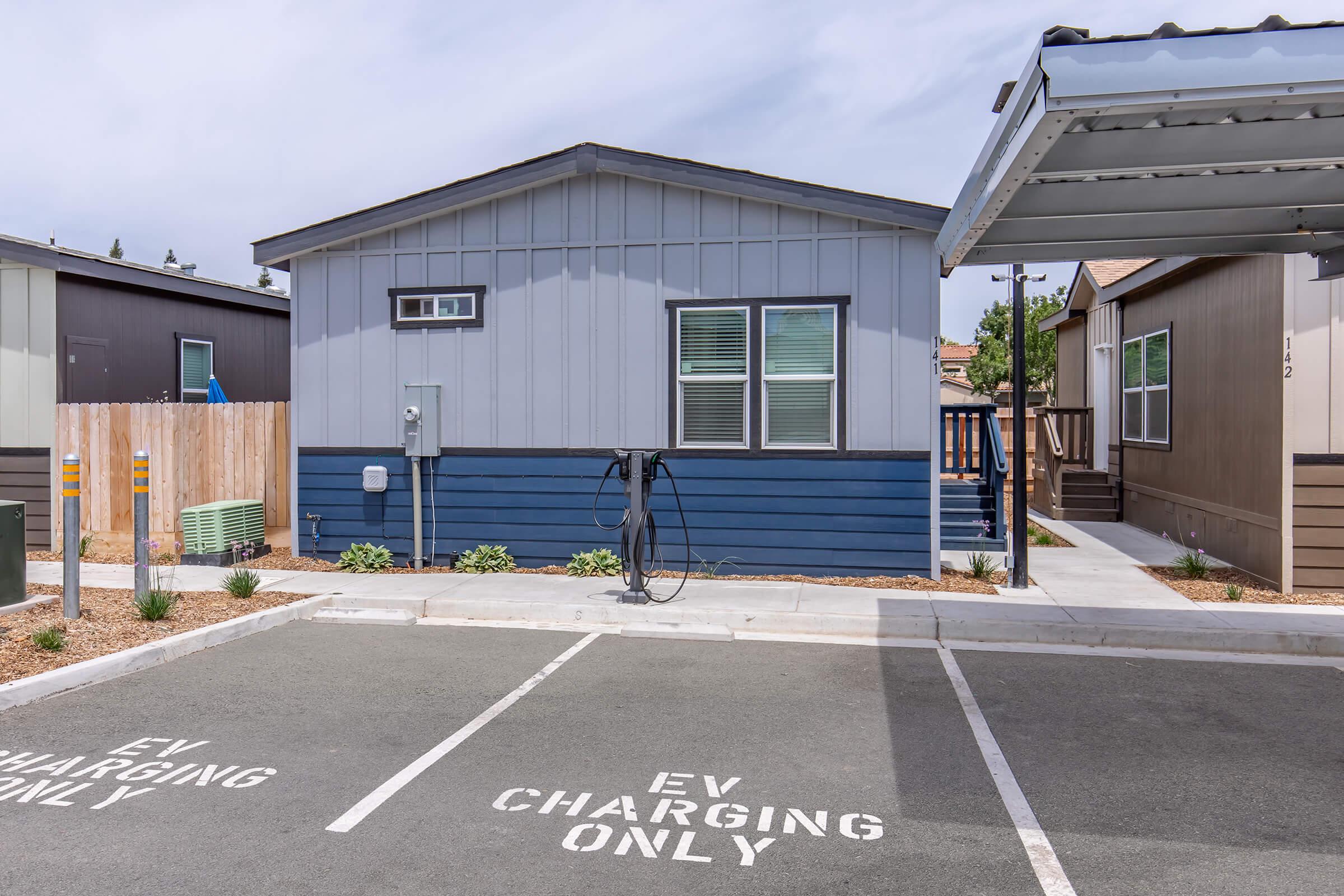 A modern residential building marked with a blue and gray exterior, featuring a driveway with "EV Charging Only" parking spaces. A charging station is visible next to the building, surrounded by landscaped areas with sparse vegetation. The setting appears to be a community with similar homes nearby.