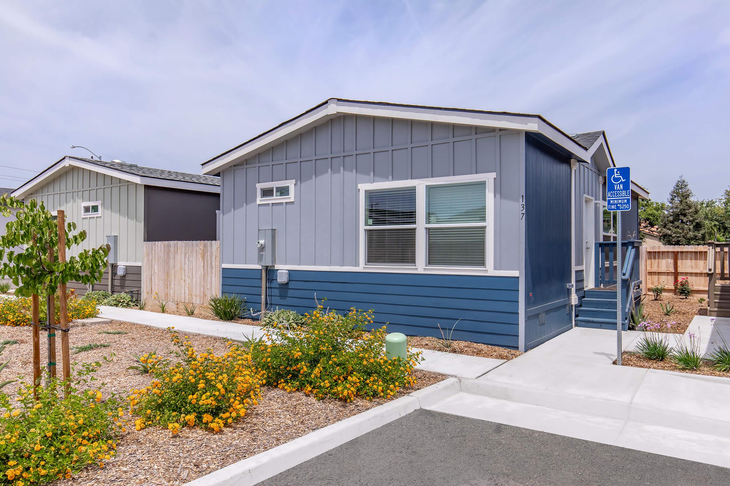 A modern, single-story house with a blue and gray exterior. The home features a front sidewalk, small garden with colorful flowers, and a wheelchair-accessible sign. The surrounding area is landscaped with gravel and low-maintenance plants, under a clear sky.