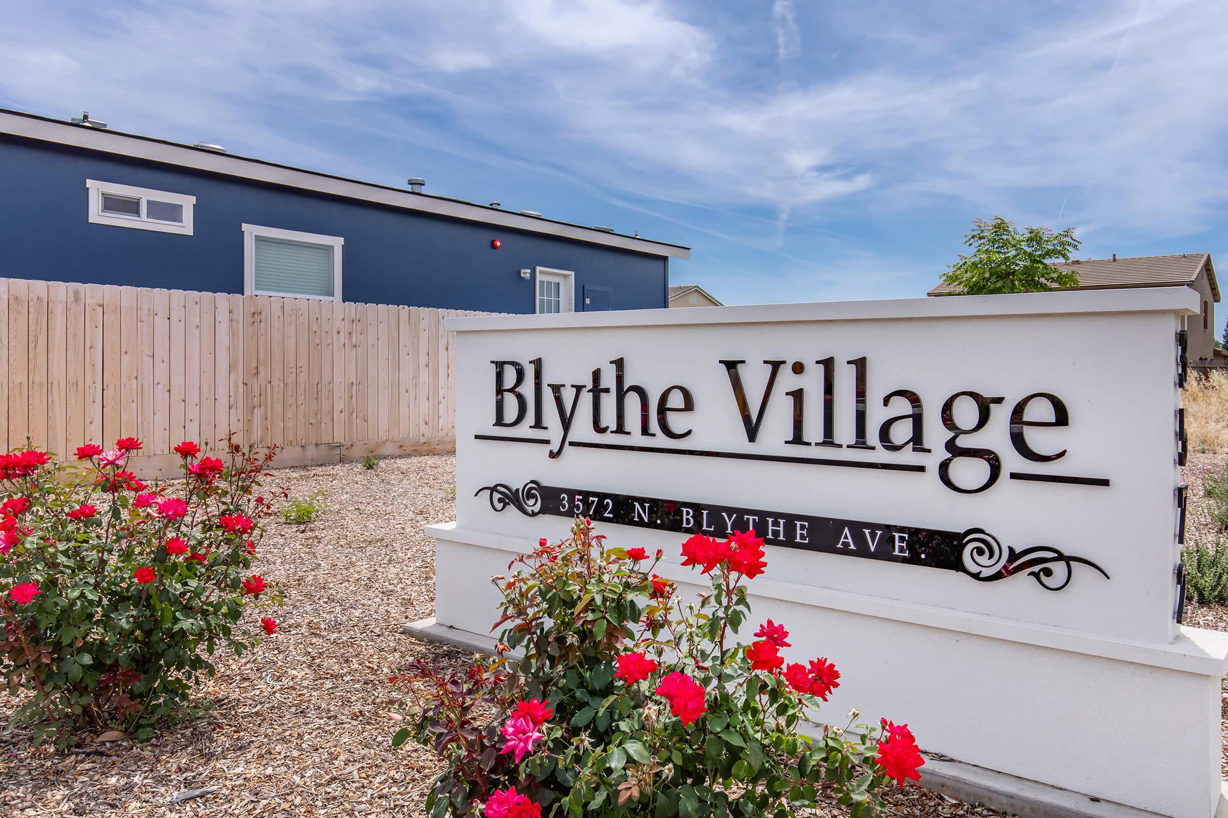 Sign for Blythe Village, prominently displaying the name "Blythe Village" in elegant lettering. The sign is surrounded by landscaped gravel with blooming red roses in the foreground. A blue building is visible in the background, along with a light-colored wooden fence under a clear blue sky.