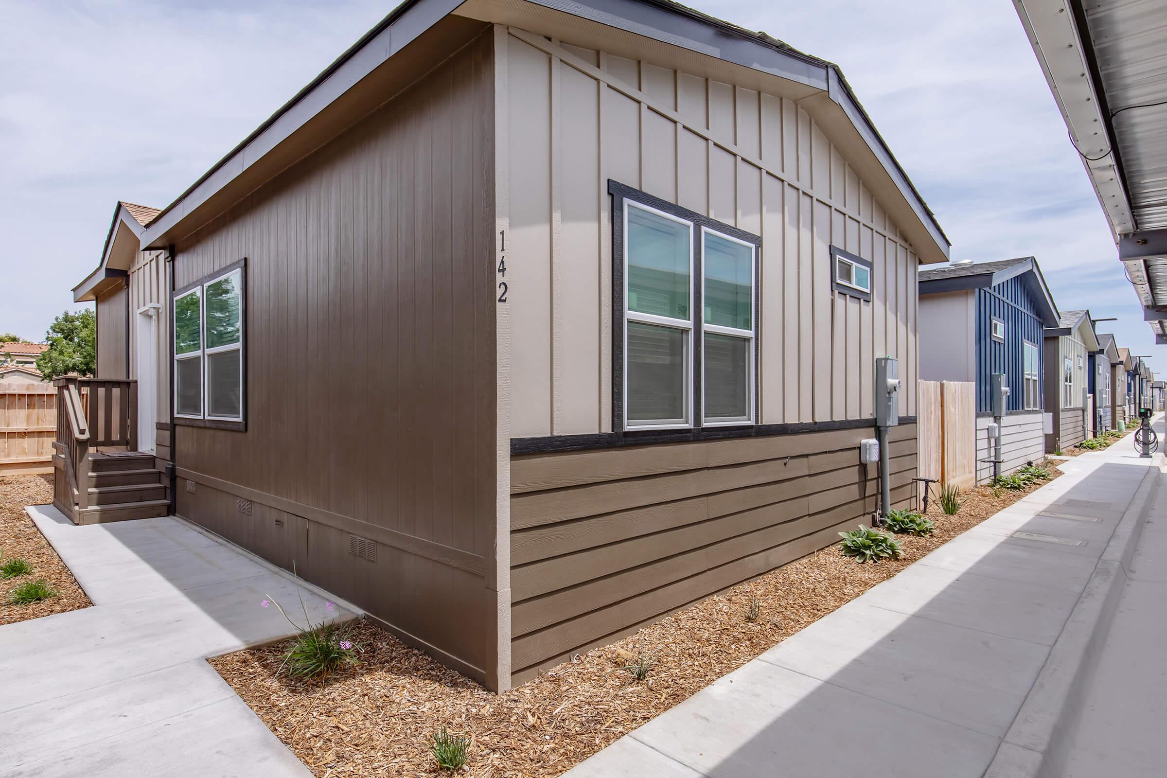 A modern, single-story house with a brown and tan exterior, featuring multiple windows and a small front porch. The house is situated between two similar homes along a clean, paved walkway lined with a small garden and new landscaping. Clear sky visible in the background.