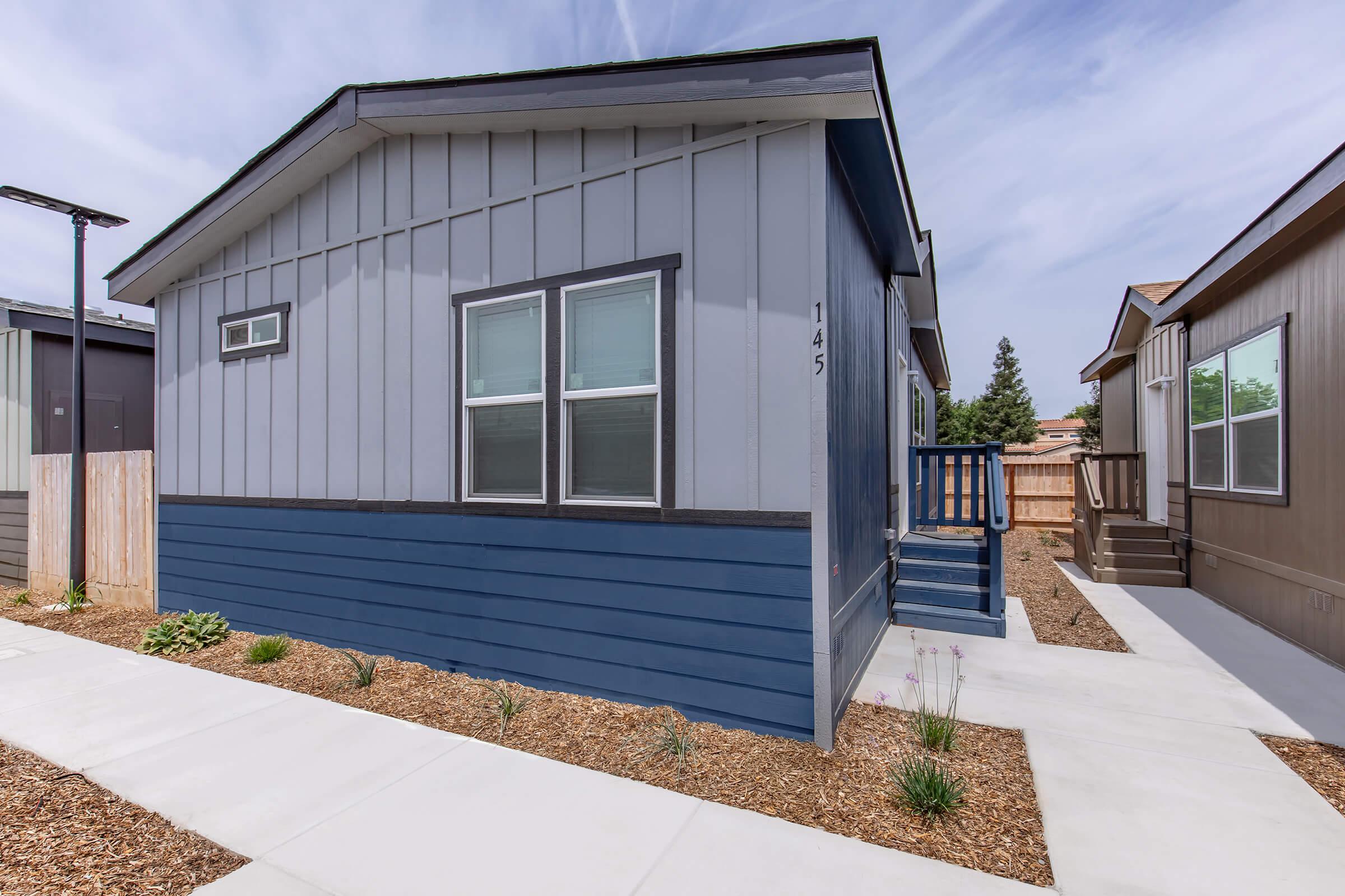 A modern single-story house with a blue and gray exterior. The house features a small porch with steps leading up to the entrance. Surrounding the house are landscaped areas with minimal vegetation and a clean concrete walkway. The sky is partly cloudy, creating a bright atmosphere.