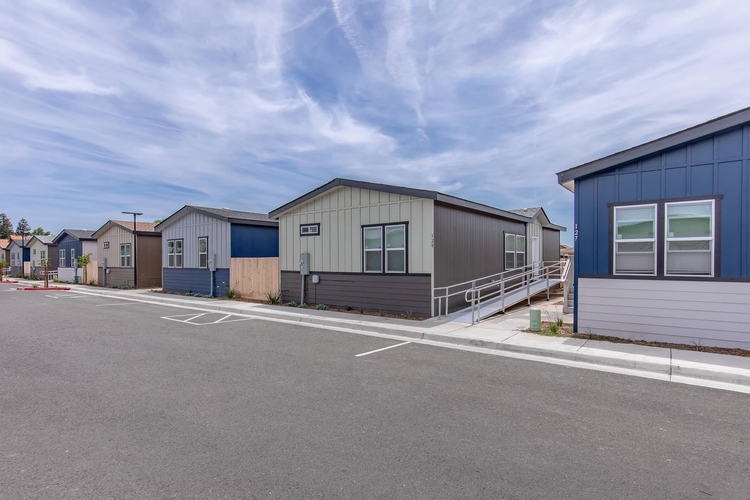 Row of modern, single-story homes in a residential area. The houses feature various siding colors, with an emphasis on gray and blue hues. Curbside parking spaces are visible. There’s a wheelchair-accessible ramp leading to one of the homes. The sky is partly cloudy, adding to the serene atmosphere.