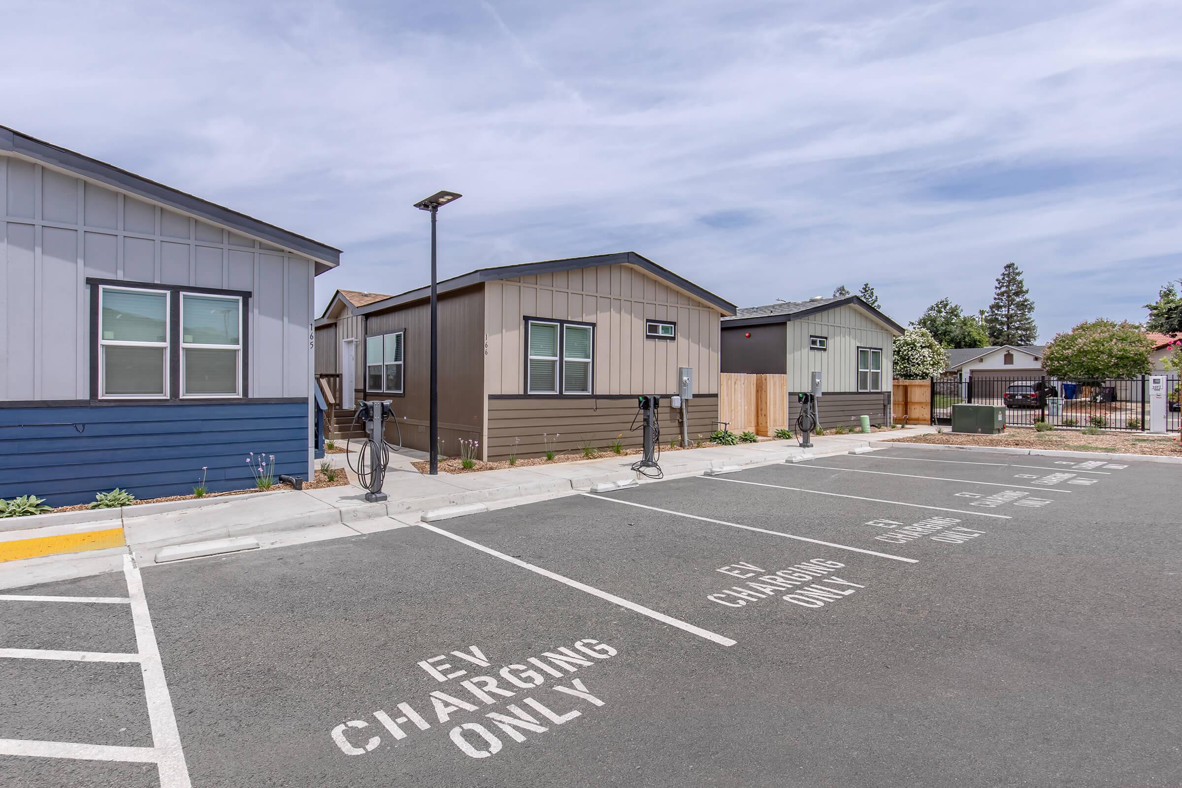 A parking lot featuring designated spaces for electric vehicle (EV) charging. Two small residential buildings are visible in the background, with a light blue building on the left and a brown building on the right. Charging stations are positioned adjacent to the parking spaces, under a clear sky.