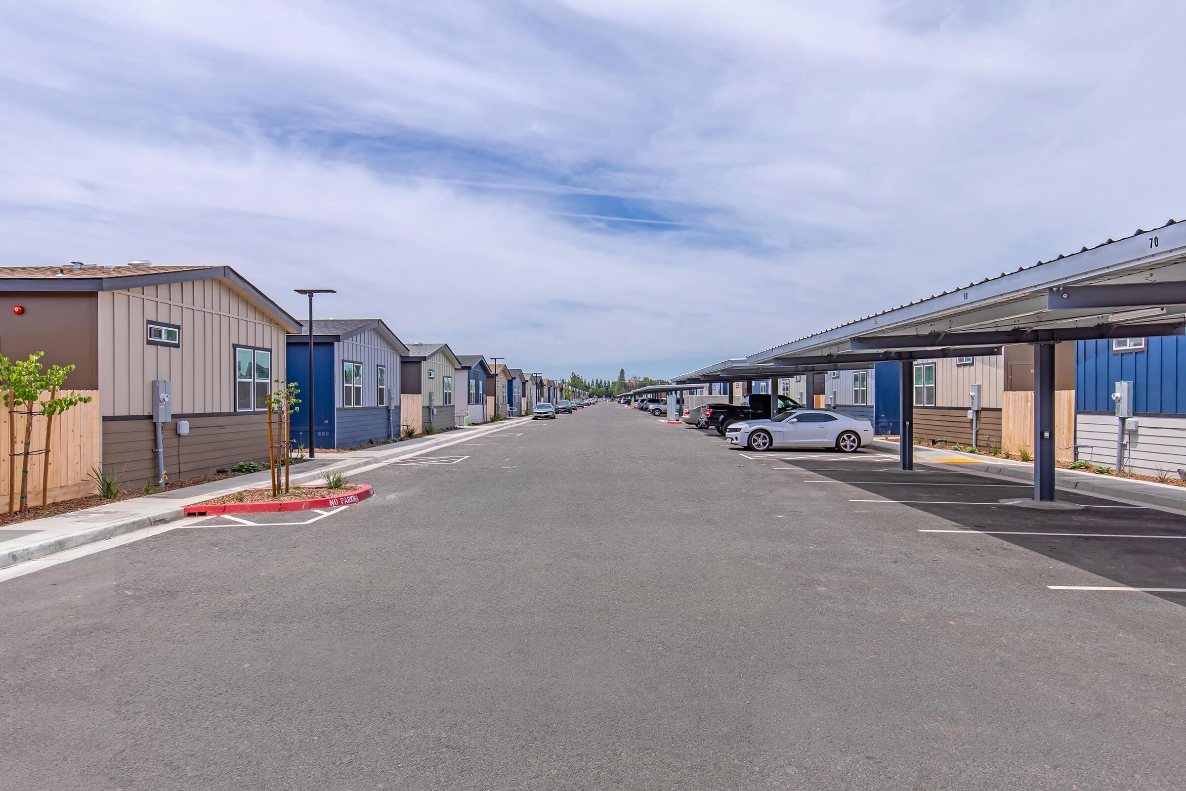 A residential street lined with modern, single-story homes on both sides. Each home features distinct colors and designs. There are covered parking spaces available, and the street is empty, with a clear sky above and a few trees lining the side. The environment appears clean and well-maintained.