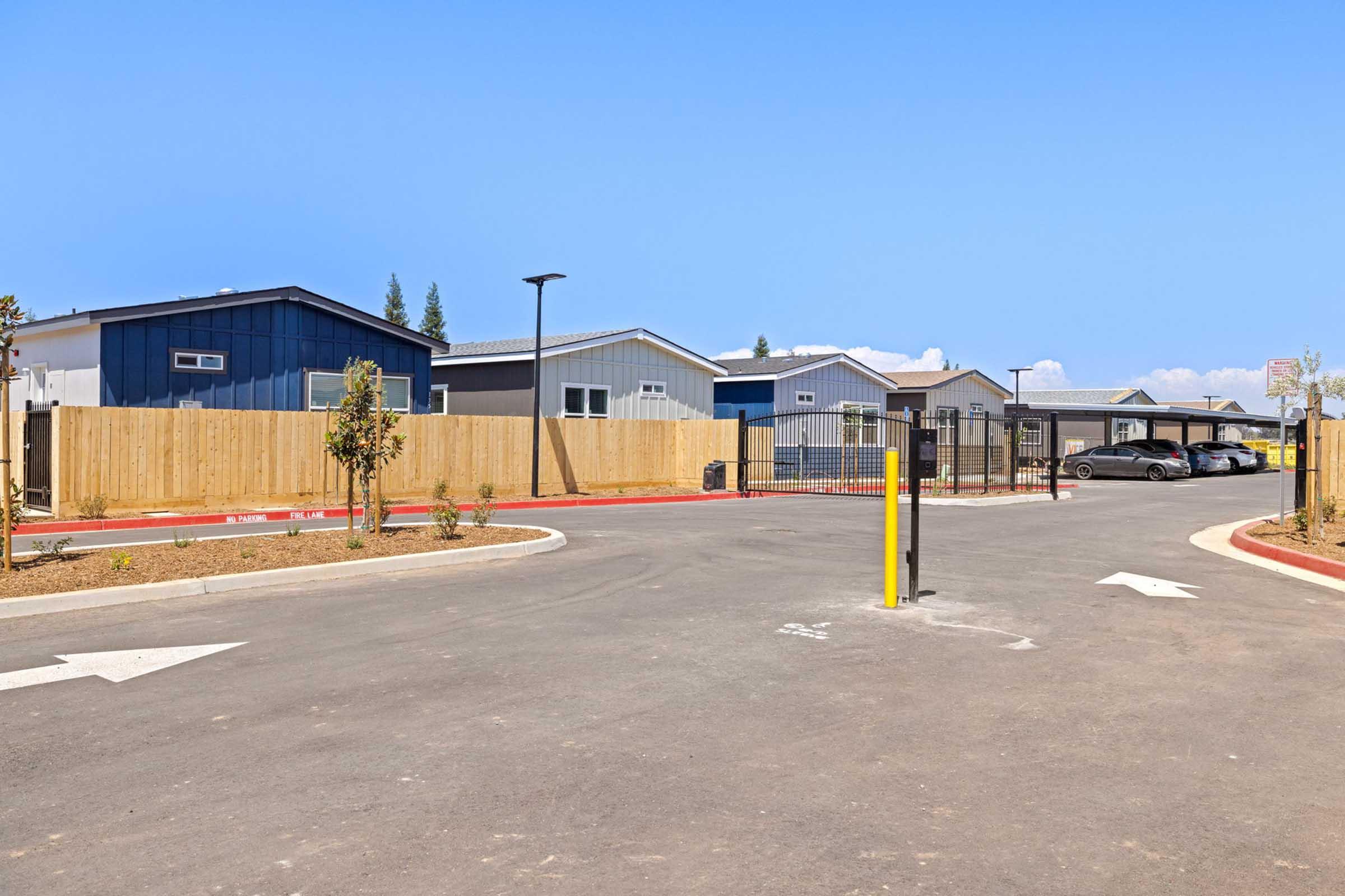 A view of a new residential development featuring several houses with a modern design. The driveway leads to a gated area with parking spaces, surrounded by wooden fencing and landscaped greenery. The sky is clear and blue, and the setting appears inviting and well-maintained.