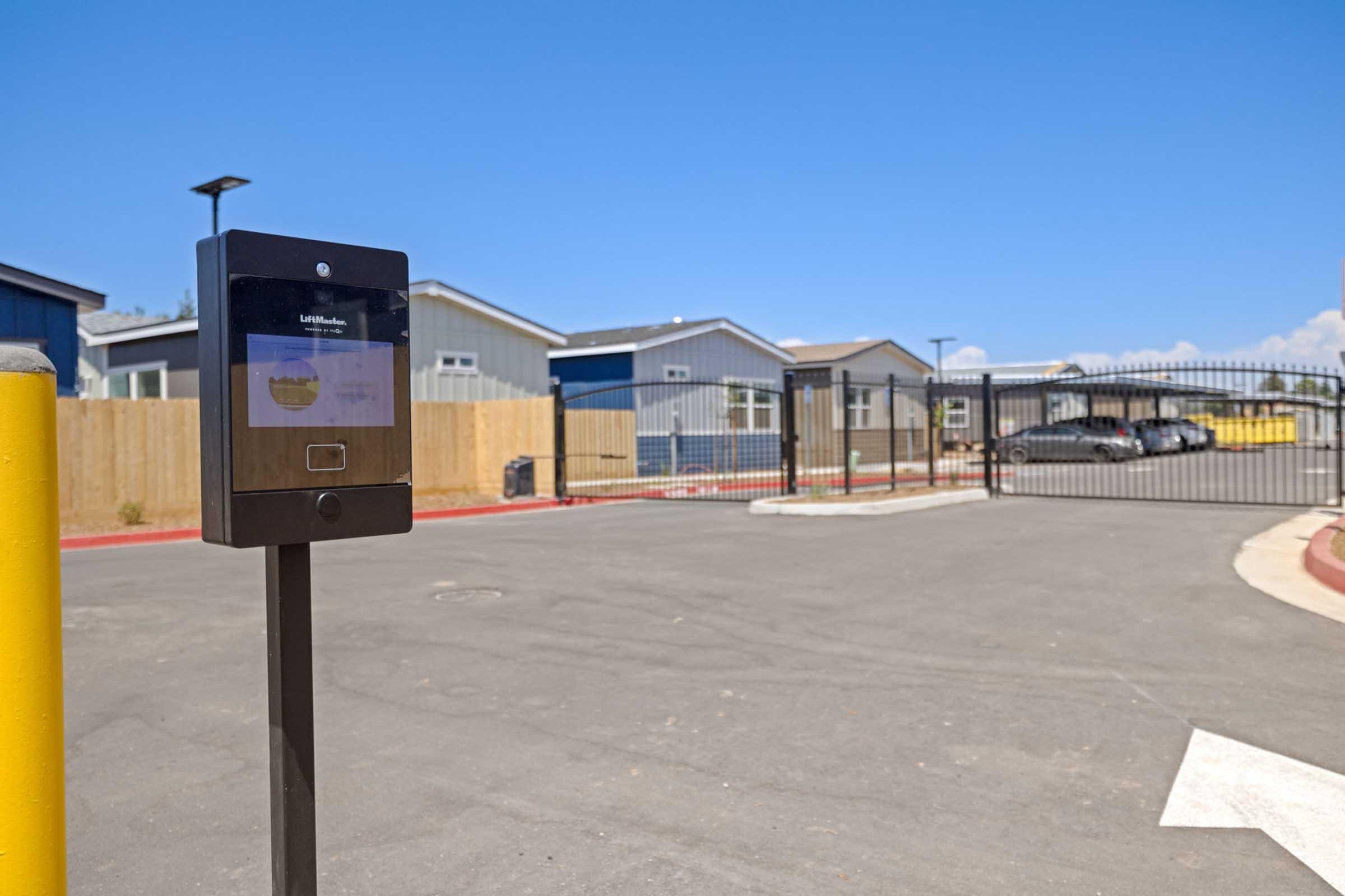 A digital access control panel is positioned next to a gated entrance. In the background, several residential buildings with varied colors are visible behind a wooden fence. The sky is clear and blue, and a parking area is seen just beyond the gate.