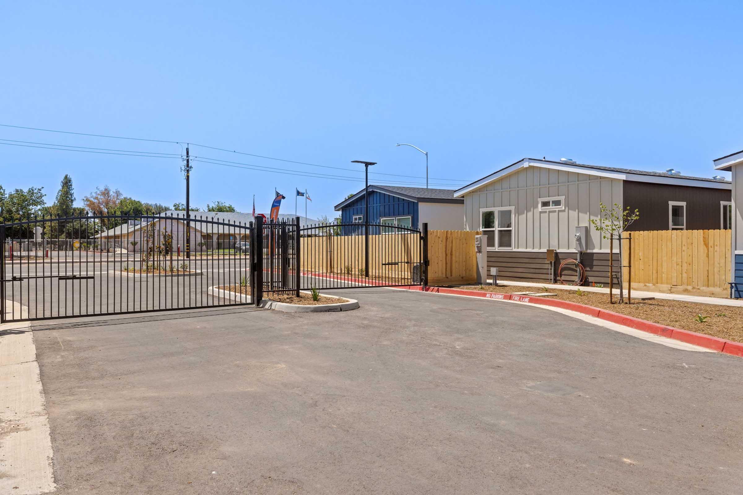 A view of an entrance gate leading to a residential area with modern buildings. The scene includes a paved road, wooden fences, and a clear blue sky. There's an American flag visible at the entrance, indicating a sense of community in the neighborhood.