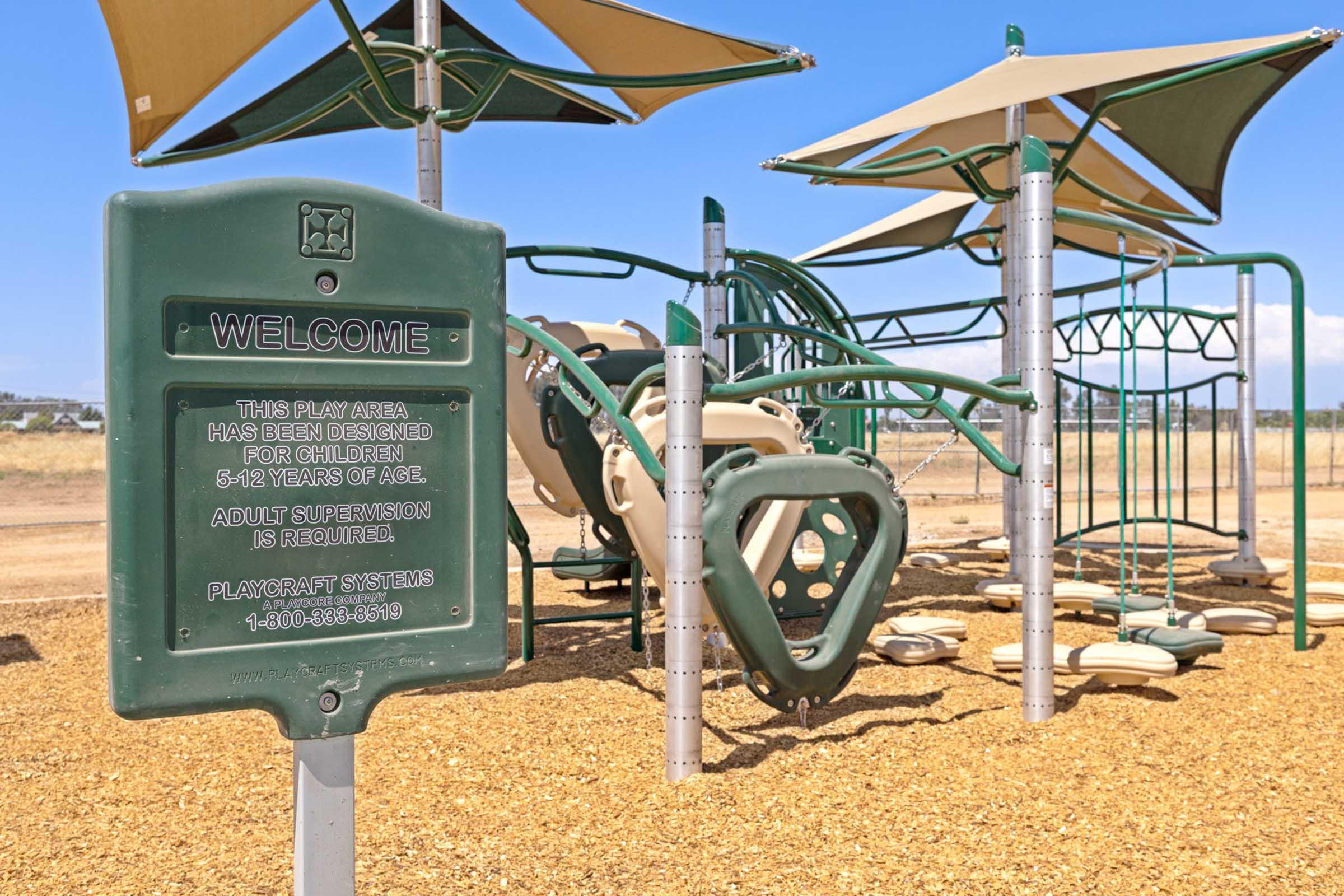 A playground area featuring climbing structures and slides, surrounded by a sandy surface. A sign indicates the space is for children aged 5-12 and emphasizes the need for adult supervision. The background shows a clear blue sky and distant landscape.